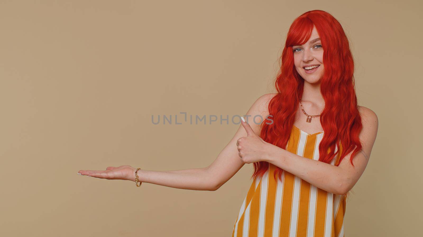 Beautiful redhead woman showing thumbs up, pointing empty place, advertising area for commercial text, copy space for goods promotion. Young ginger girl. Studio shot indoors on beige wall background