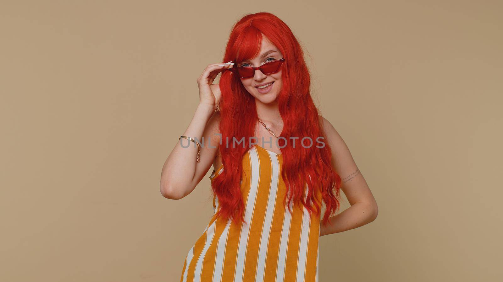 Portrait of seductive cheerful stylish woman in tank top wearing sunglasses, charming smile. People emotions concept. Young lovely redhead ginger girl posing isolated alone on beige studio background