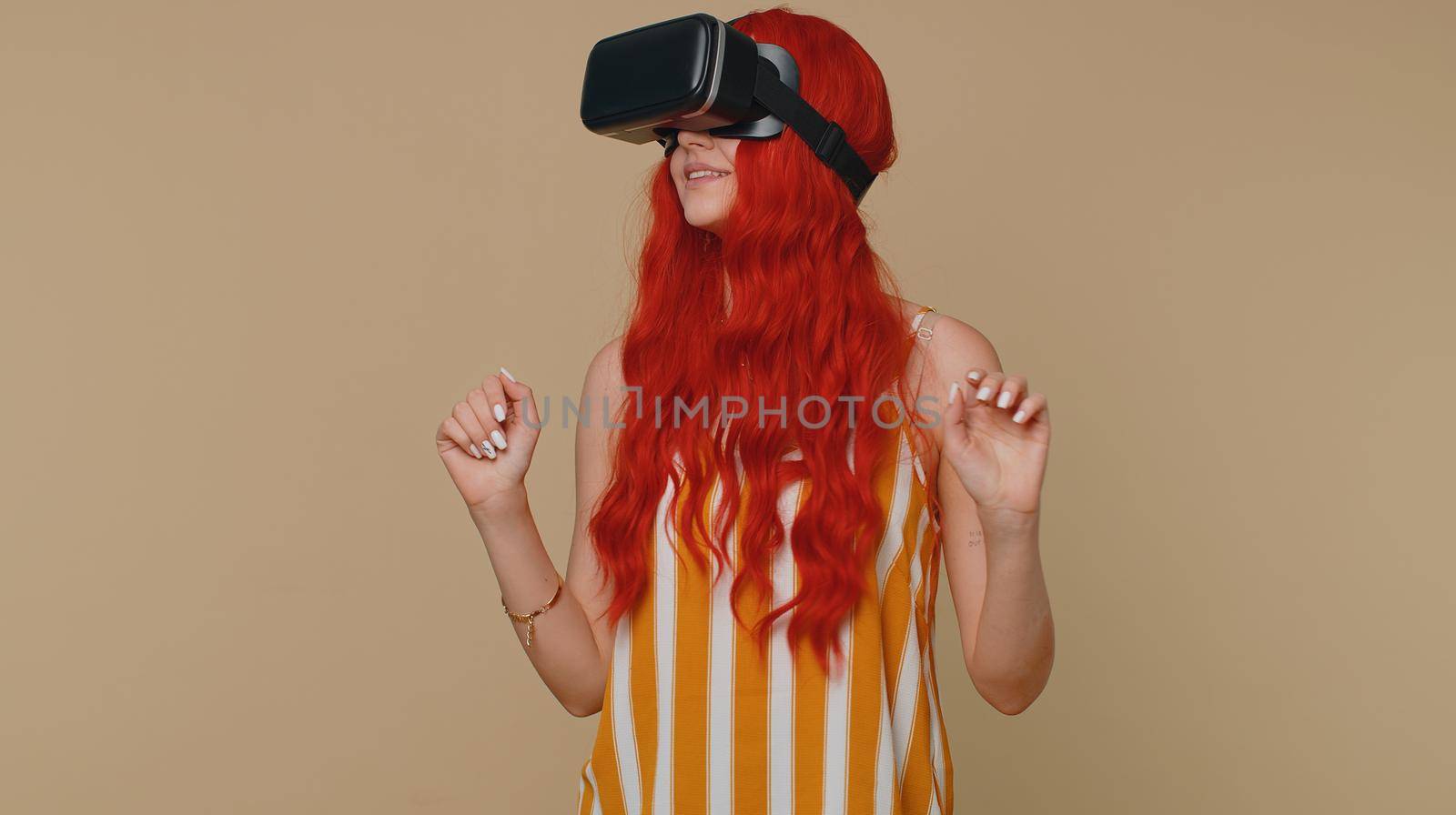 Exited redhead woman using virtual reality futuristic technology VR app headset helmet to play simulation 3D 360 video game, drawing. Ginger girl on beige studio background. People sincere emotions