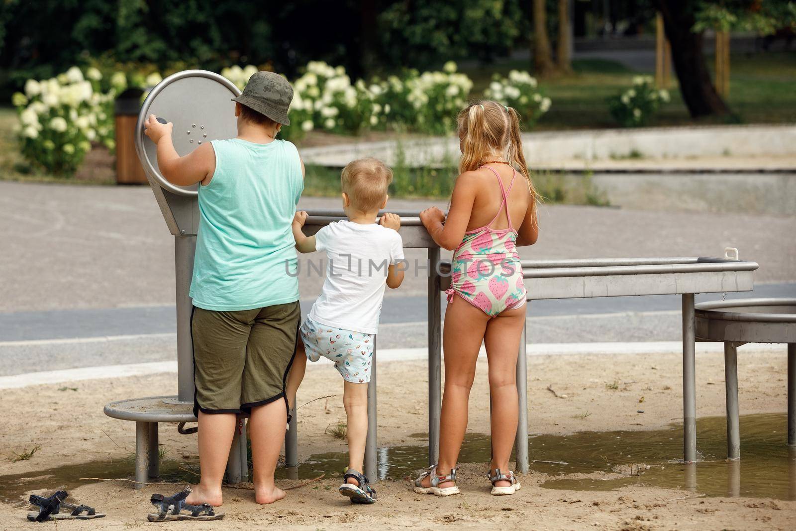 Little boy wearing shorts and white t-shirt playing with water from public faucet, his friends big boy and girl pumping.