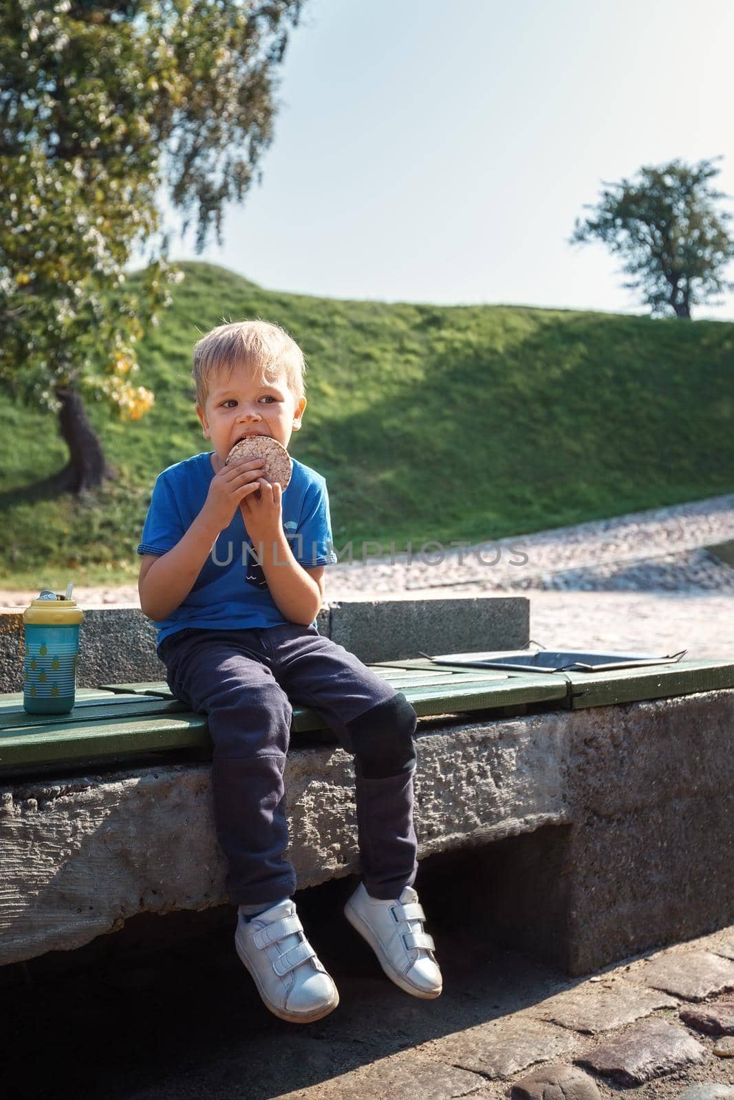Adorable baby boy eat biscuit or cookie sitting on wooden bench at outdoor park. Happy kid enjoying with food, he licks the chocolate and smears his nose.