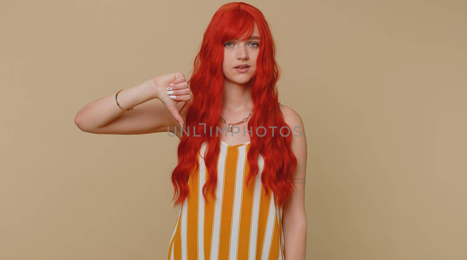 Upset unhappy red hair woman in striped tank top showing thumbs down sign gesture, expressing discontent, disapproval, dissatisfied, dislike. Young ginger girl. Indoor studio shot on beige background