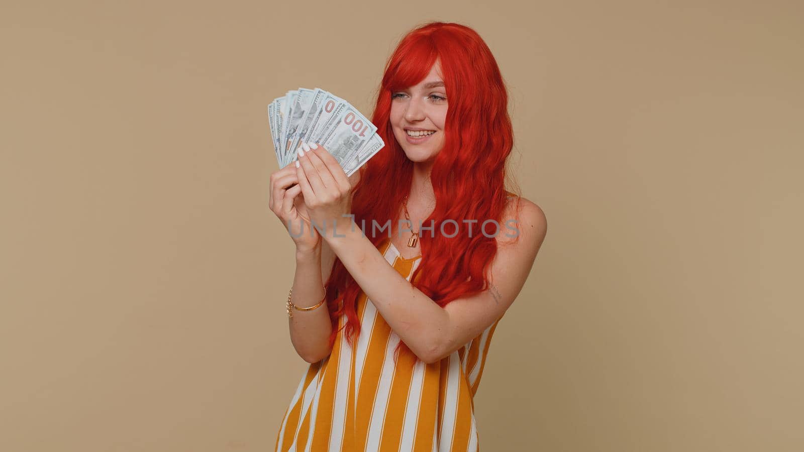 Redhead young woman holding fan of cash money dollar banknotes celebrate dance, success business career, lottery game winner, big income, wealth. Red hair ginger girl alone on beige studio background