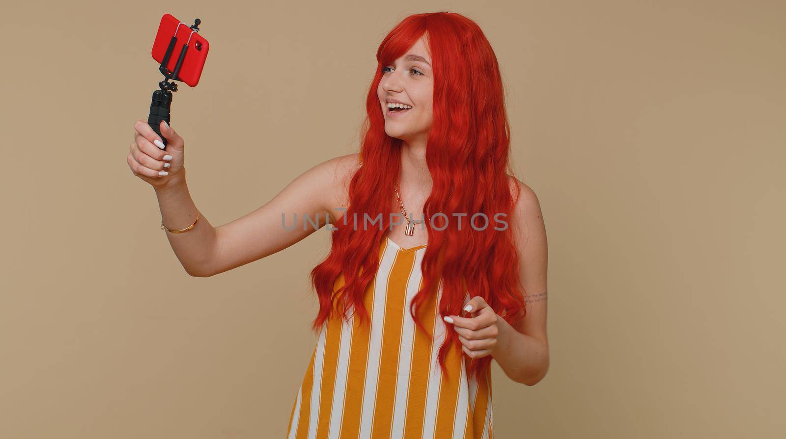 Redhead woman in tank top traveler blogger taking selfie on mobile phone selfie stick, communicating video call online with subscribers. Young ginger girl isolated alone on beige studio background
