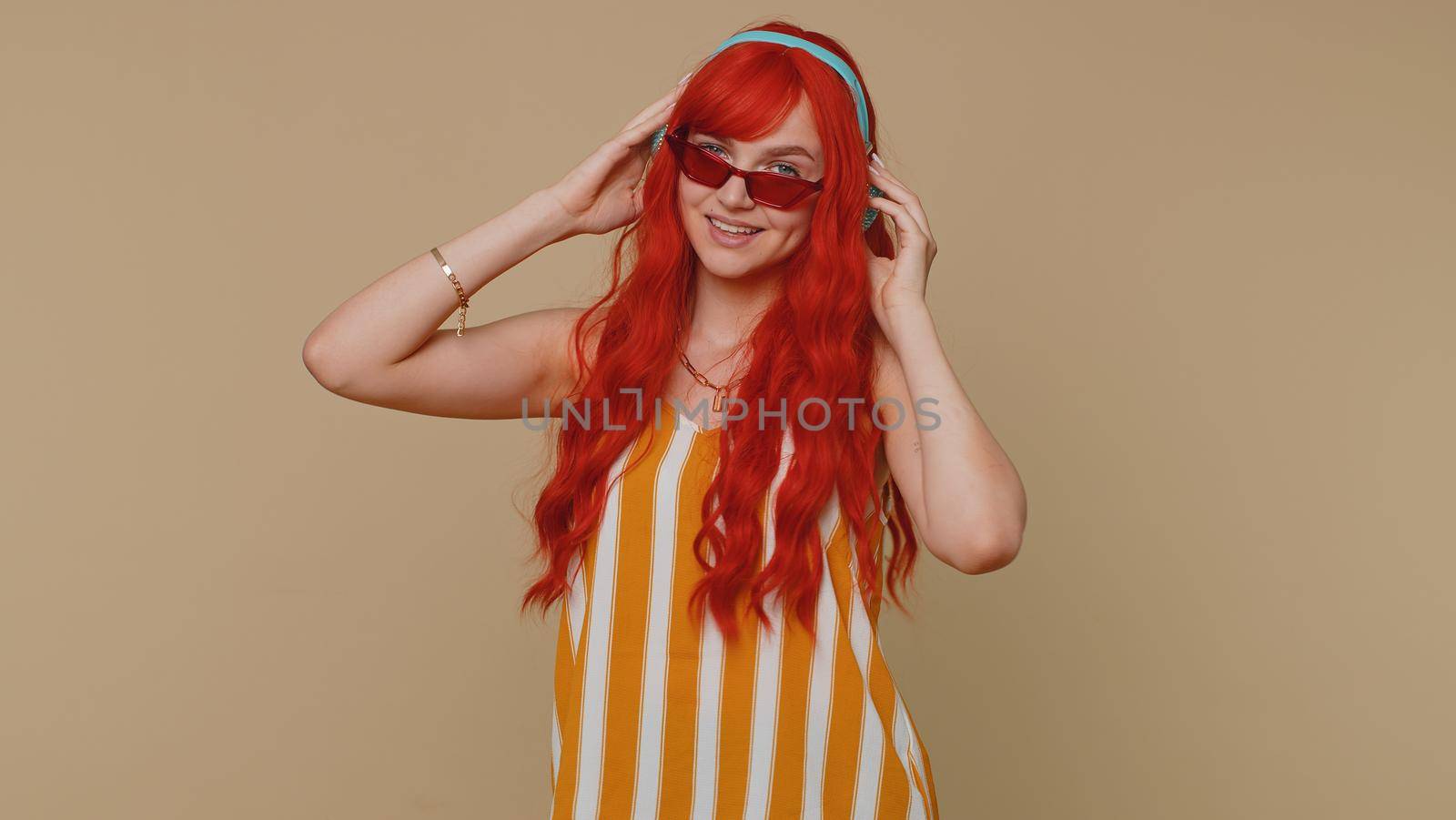 Portrait of redhead woman in tank top listening music via headphones and dancing disco fooling around having fun expressive gesticulating hands. Young one ginger girl on beige studio wall background