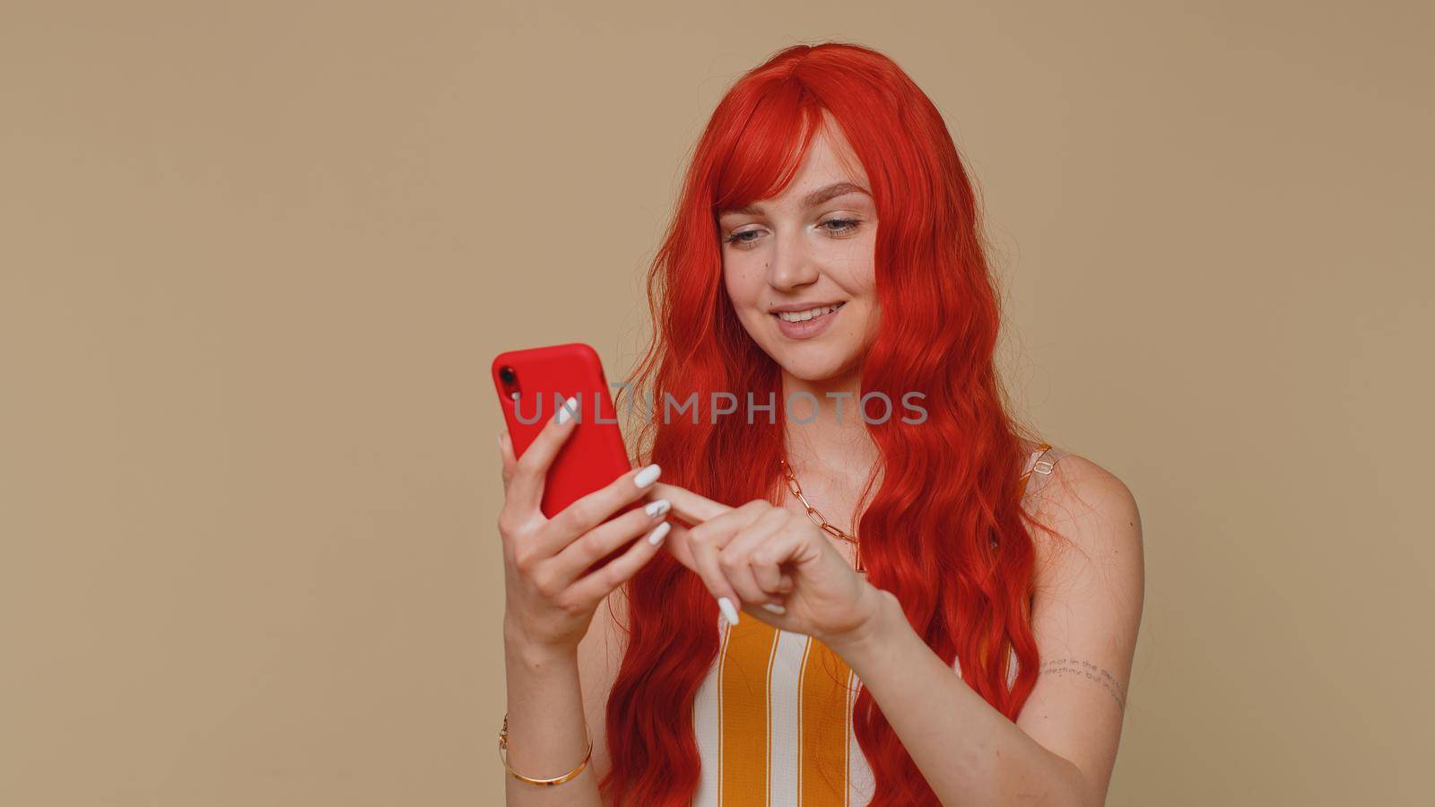 Beautiful red hair woman using mobile phone typing new post on web sms message, browsing, searching addiction of social networks. Young ginger girl studio shot isolated alone on beige wall background
