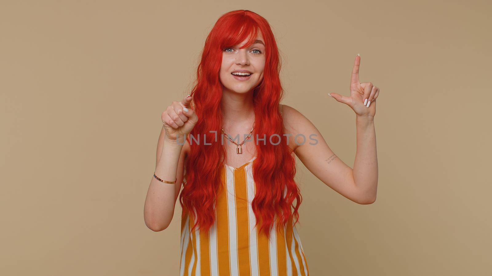 Woman showing loser gesture and pointing on you, blaming accusing for unsuccess, expressing disrespect, mocking your failure, bullying abuse. Young redhead girl isolated on beige studio background