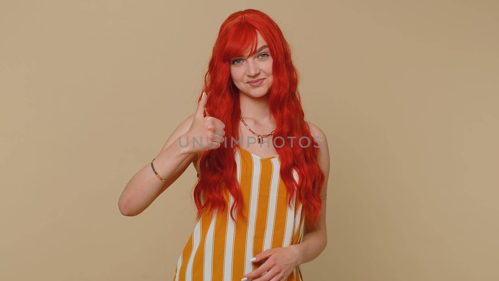 Redhead young woman in striped tank top raises thumbs up agrees with something or gives positive reply recommends advertisement likes good. Adult ginger girl isolated alone on beige studio background
