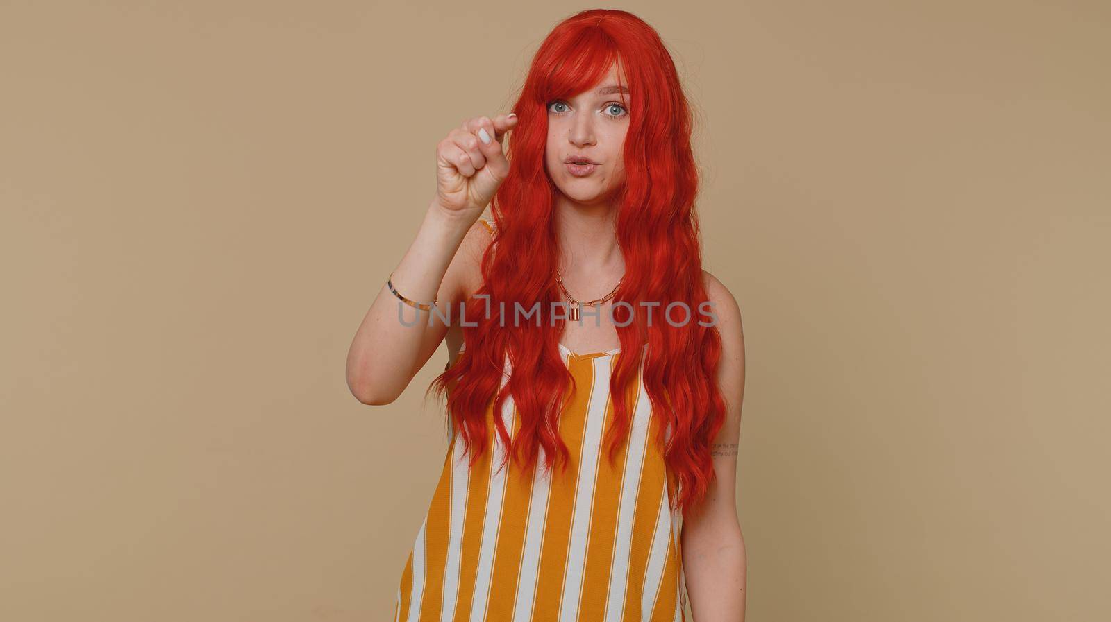 Hey you. Ginger woman in tank top smiling excitedly and pointing to camera, choosing lucky lottery winner, indicating to awesome you. Young redhead girl isolated alone on beige studio wall background