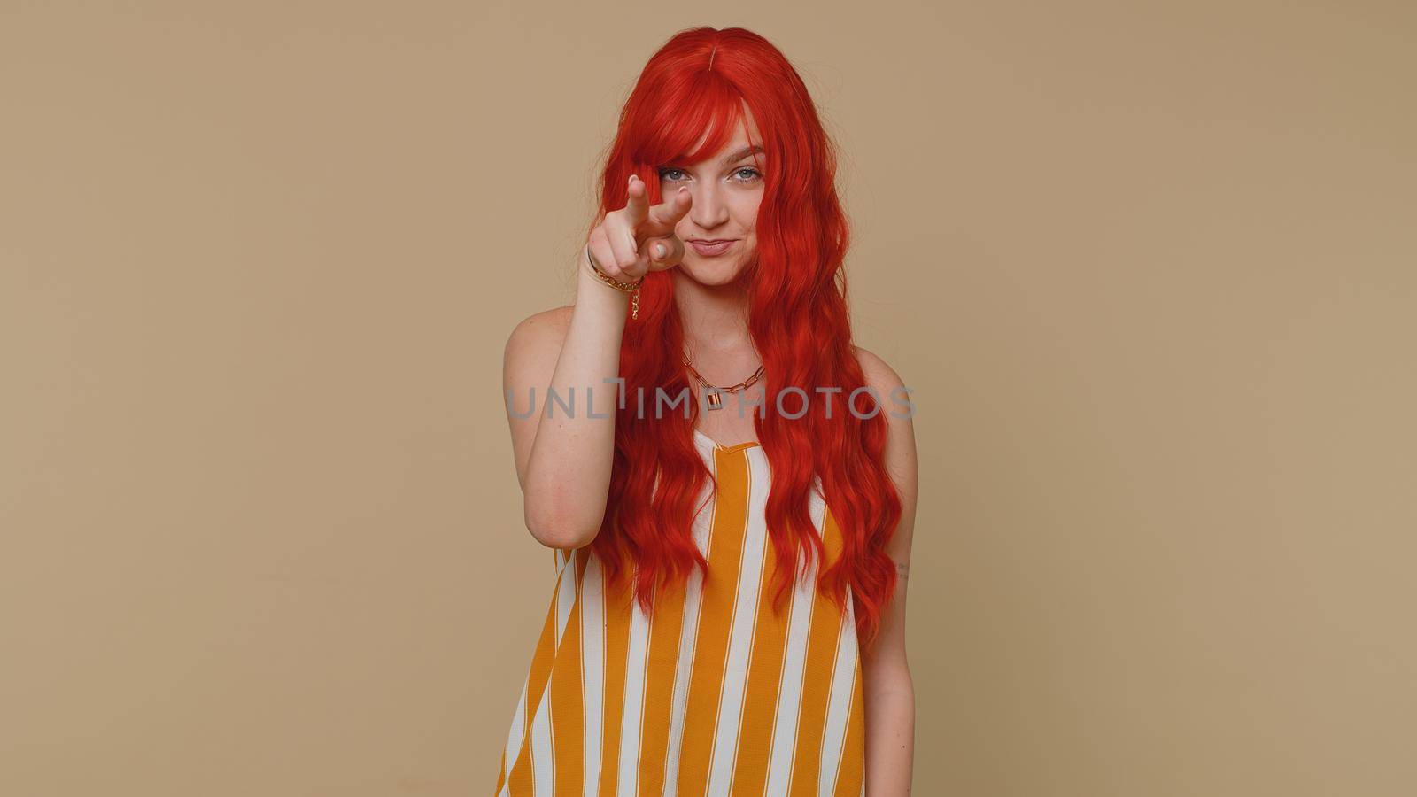 I am watching you. Ginger woman in tank top pointing at her eyes and camera, show I am watching you gesture, spying on someone. Young adult red hair girl isolated alone on beige studio wall background