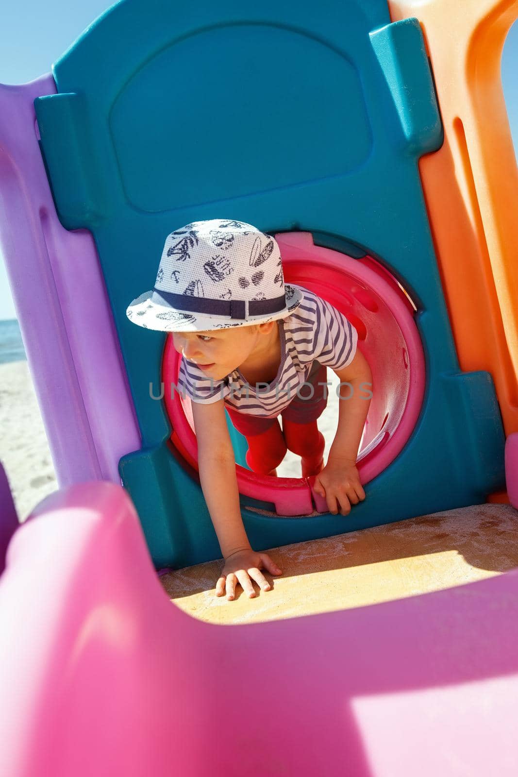 A child plays on a colourful, plastic beach playground, he emerges from a tunnel