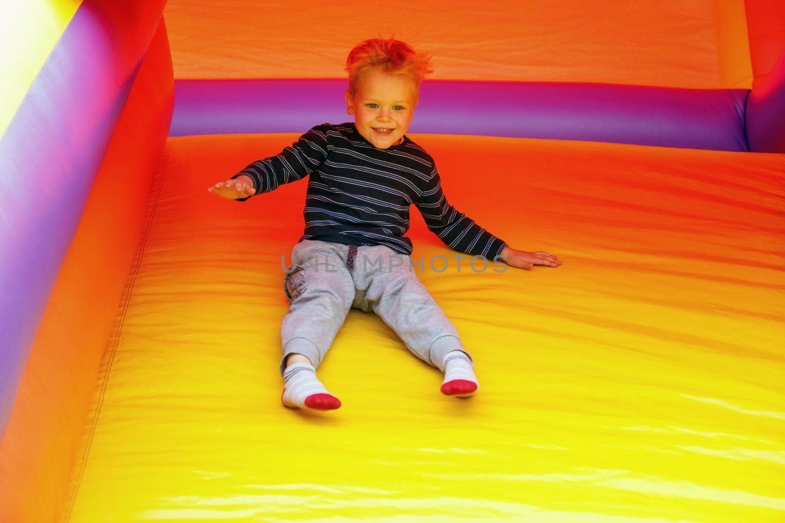 A cheerful, laughing boy sitting and slide down on a bright yellow rubber trampoline. Image in motion, a brave child is not afraid of anything by Lincikas