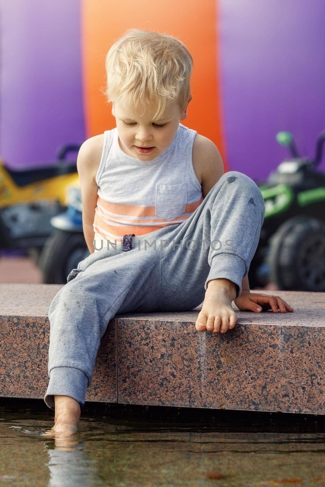 The little cute boy sits on the parapet of the fountain, he dips one foot into the water and checks the temperature in the pool.