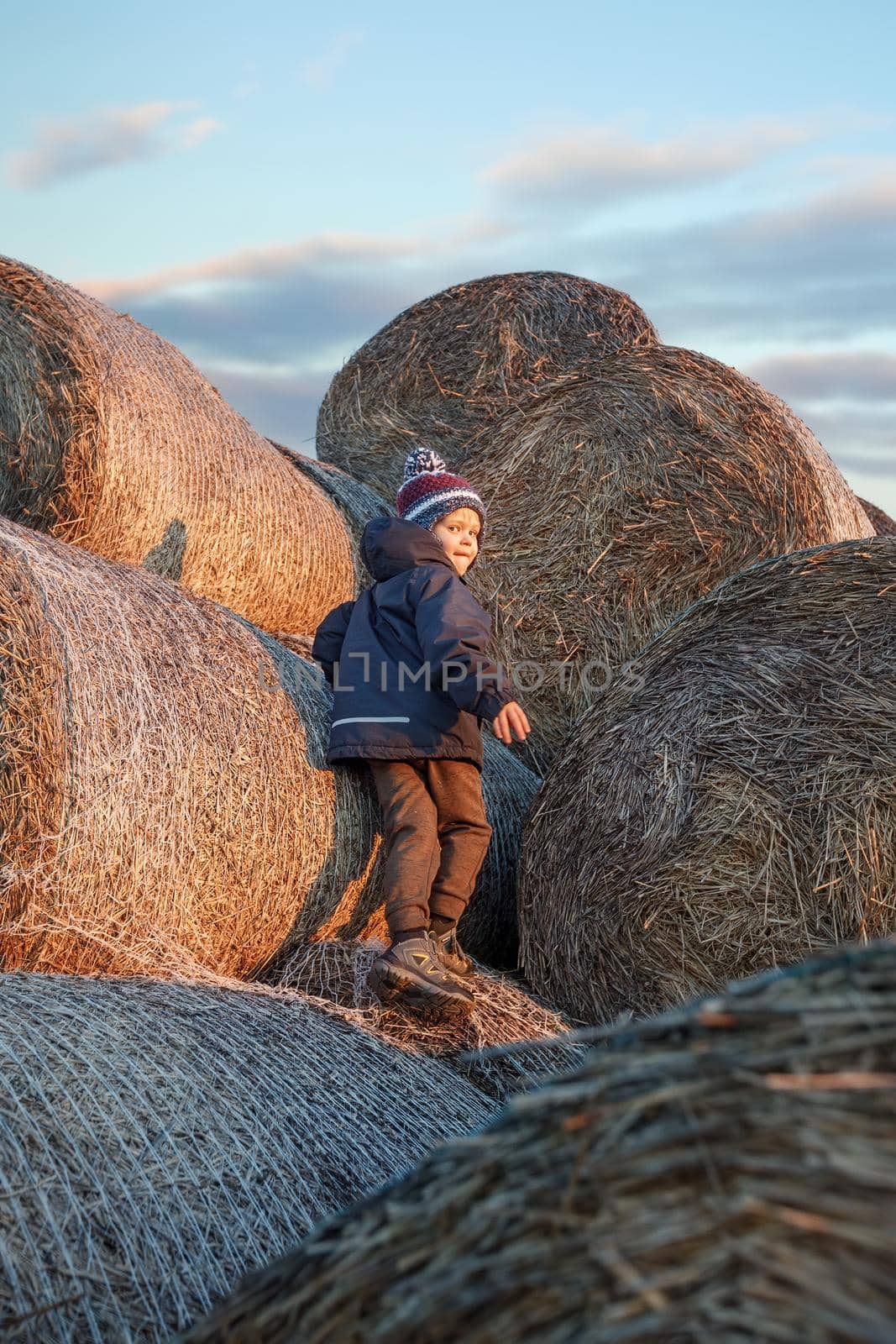 The little boy, wearing autumn clothes and a grandmother's knitted hat, plays outside on a pile of soft straw bales on an autumn evening.