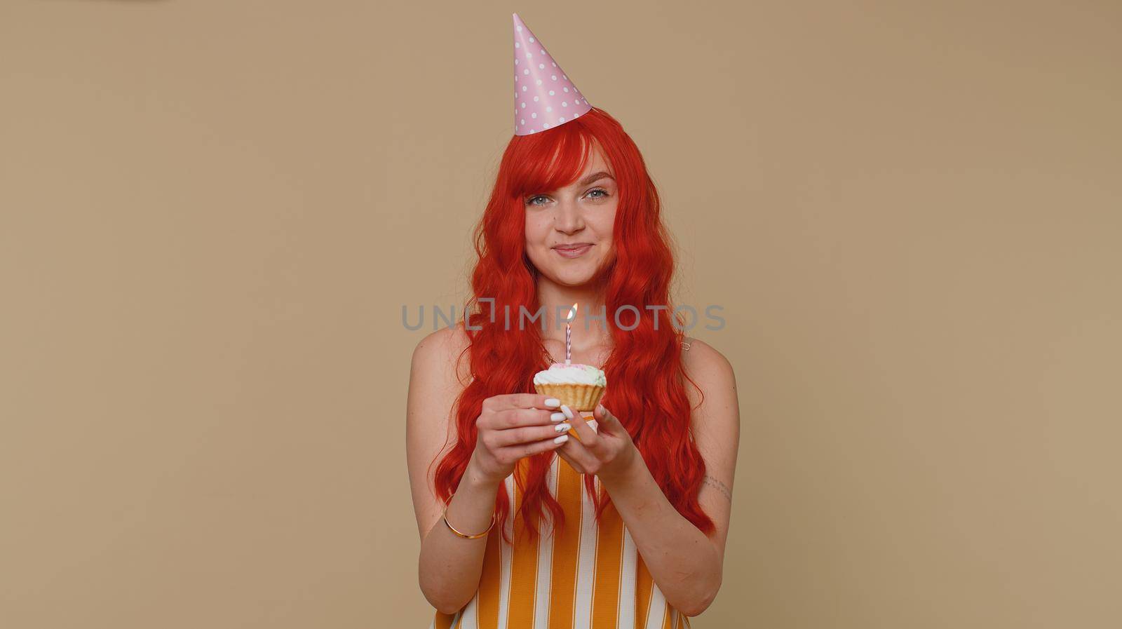 Happy redhead woman in festive cone cap celebrating birthday anniversary party, blowing candle on small cake cupcake making a wish. Red hair ginger adult girl isolated alone on beige studio background