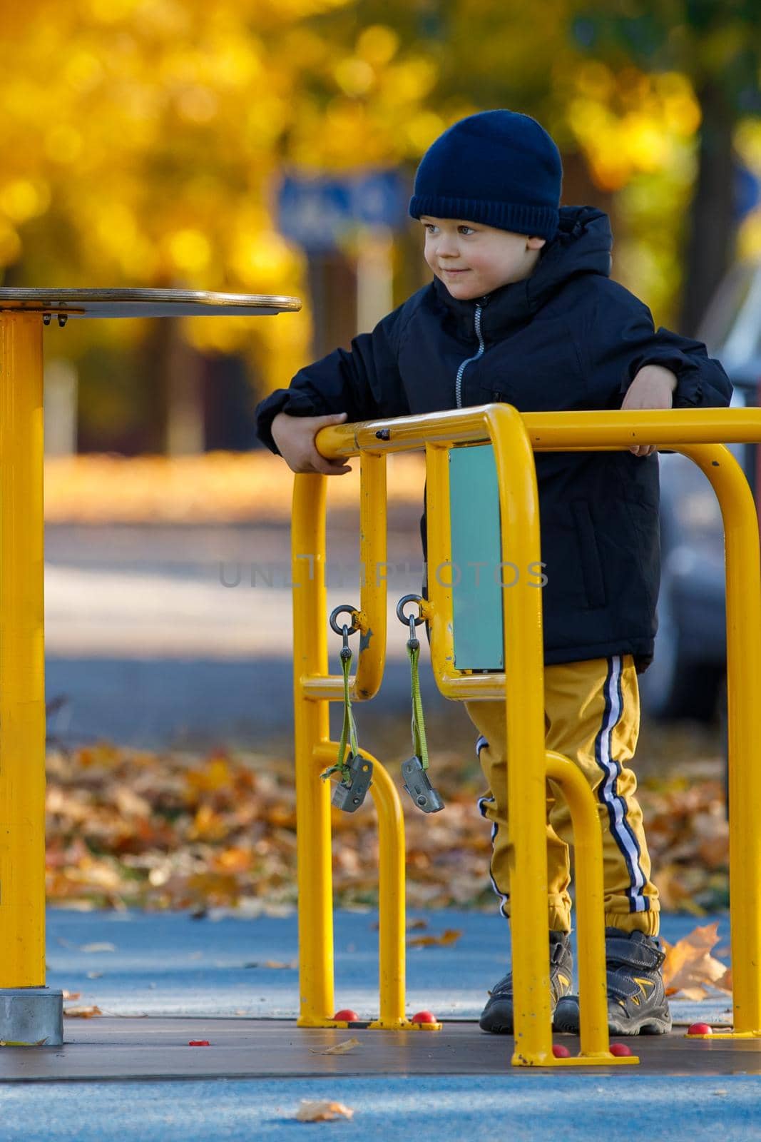 Little boy play on the playground in the park. Child spinning a yellow carousel. Autumn season with bright yellow trees in the background. Vertical photo. by Lincikas