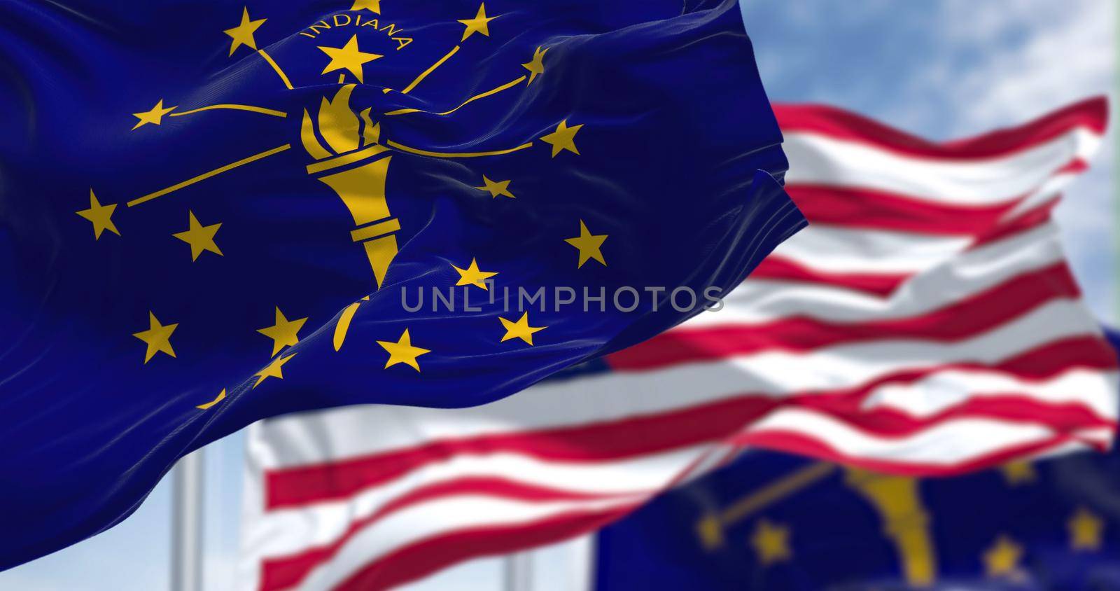 The Indiana state flag waving along with the national flag of the United States of America. In the background there is a clear sky. Indiana is a U.S. state in the Midwestern United States