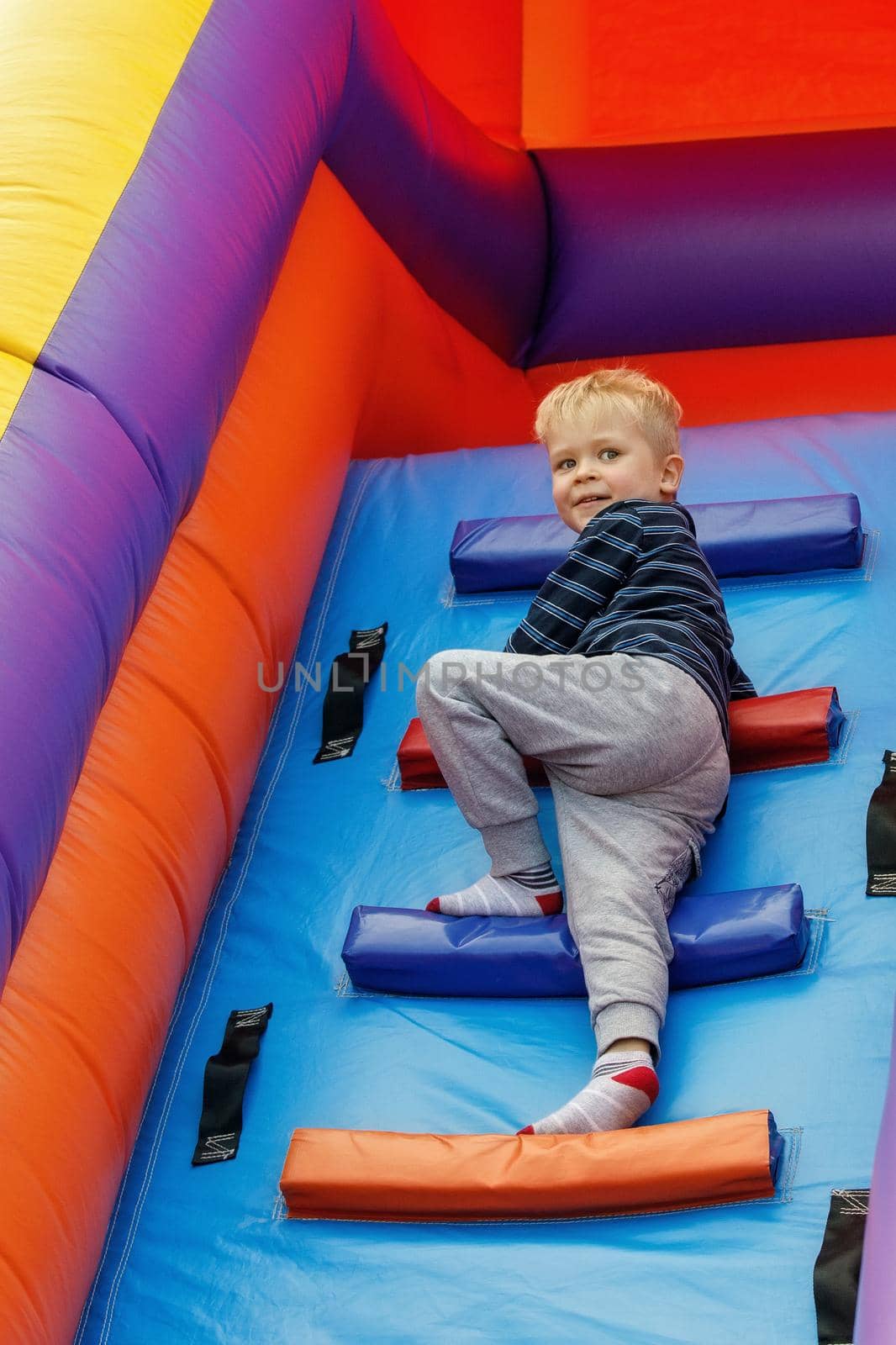 A cheerful smiling boy sports in the amusement park climbs the high stairs of an inflatable trampoline.