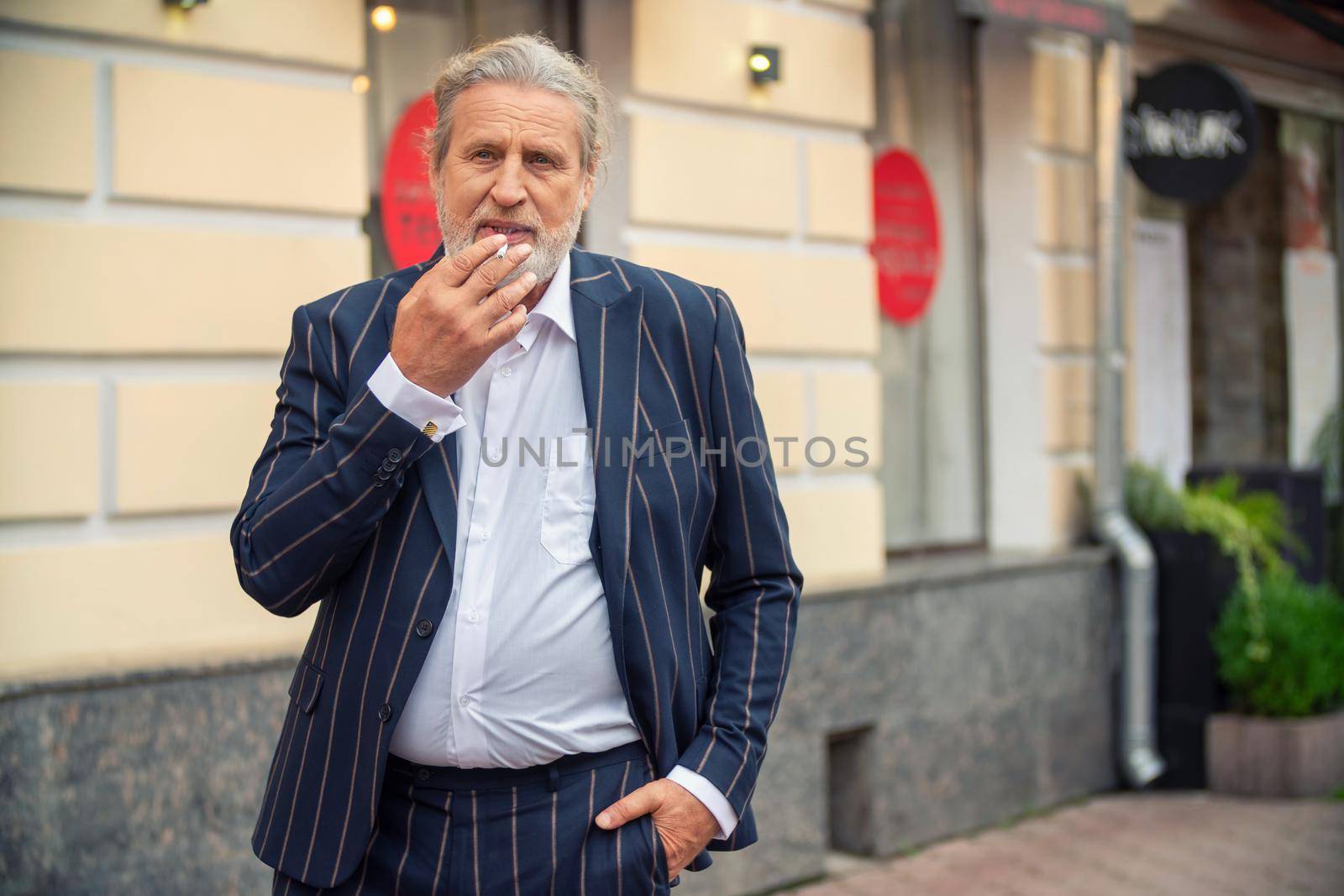 an elderly man smokes in the street with one hand in his pants pocket
