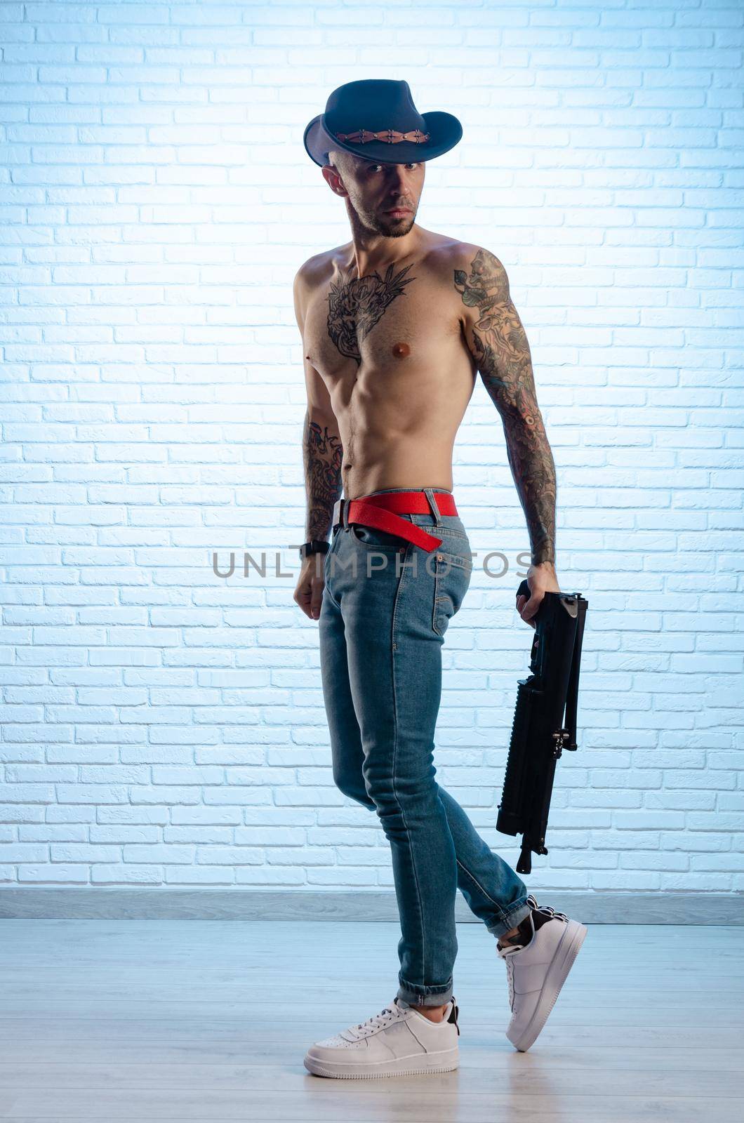 fashionable slim guy with a bare torso in tattoos, jeans and a cowboy hat with a gun by Rotozey