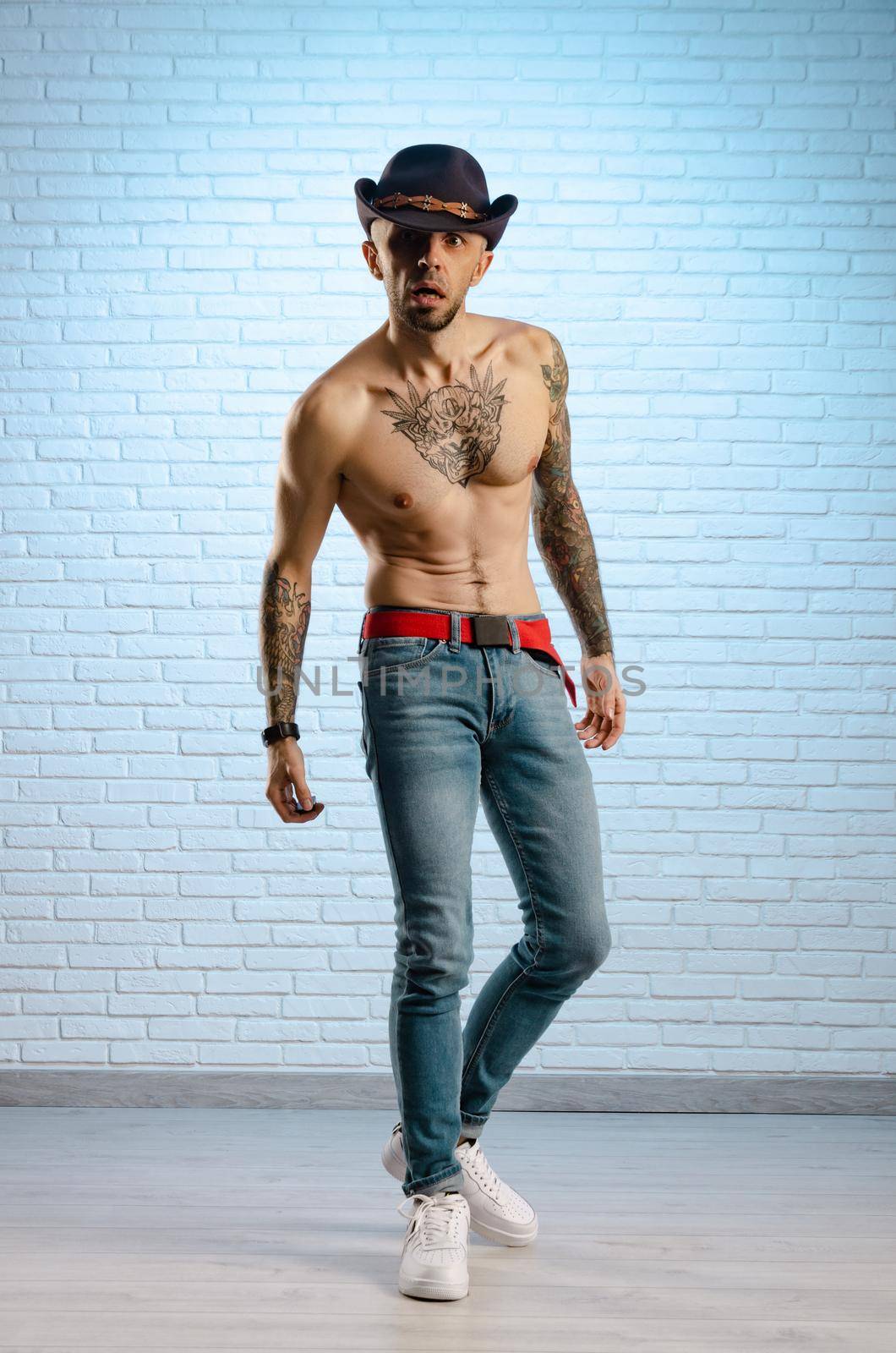 fashionable slim guy with a bare torso in tattoos, jeans and a cowboy hat by Rotozey