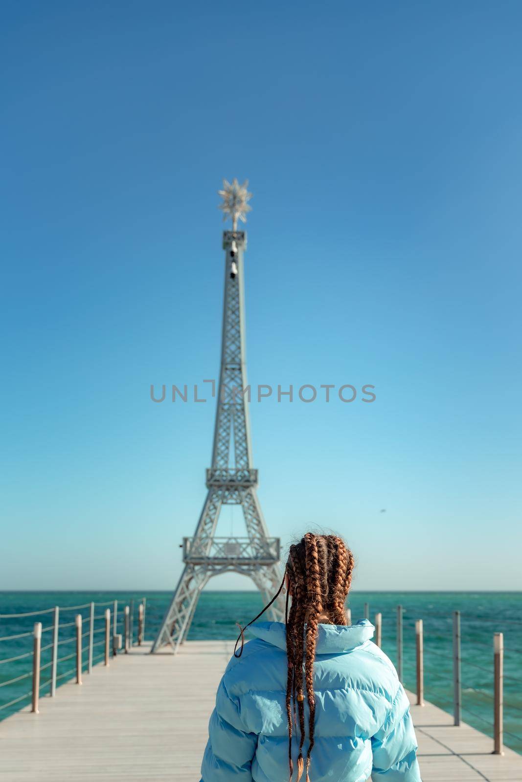 Large model of the Eiffel Tower on the beach. A woman walks along the pier towards the tower, wearing a blue jacket and white jeans