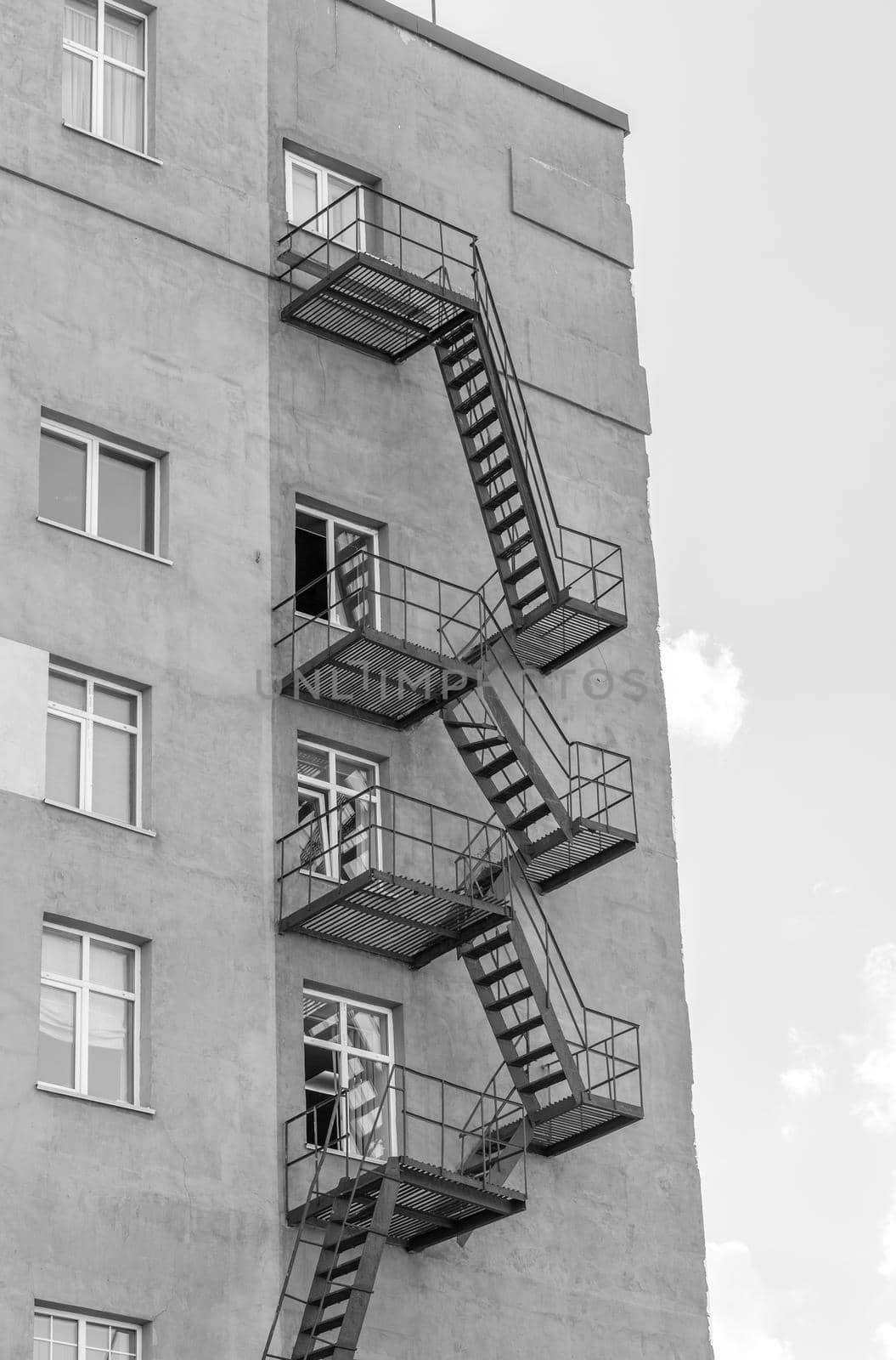 Silhouette of a fire escape on a high-rise building against a blue sky with clouds. Some of the stairs are broken. There is free space for text. Black and white photo