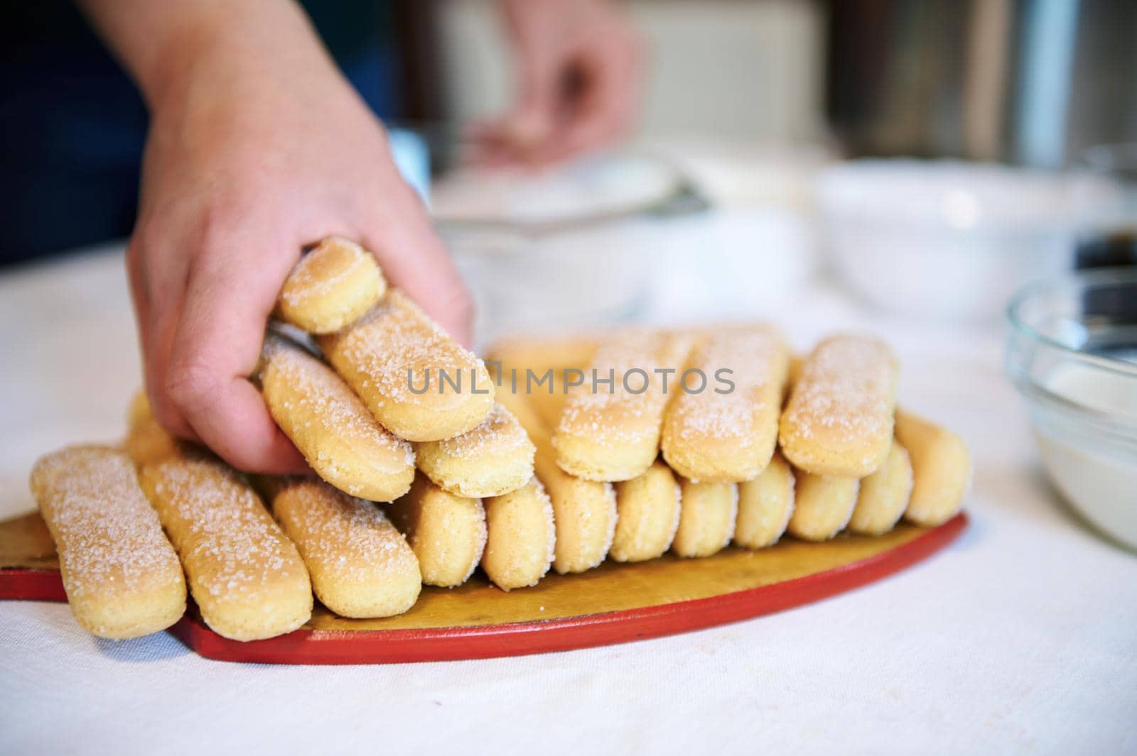 Details of a human hand holding savoiardi biscuit cookies staked on a wooden board. Cooking at home, home food and culinary concept.