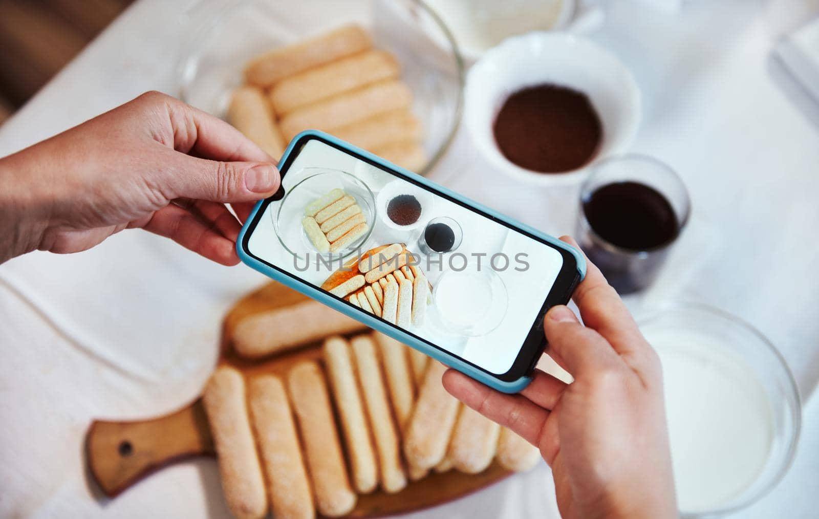 View from above. Hands using smartphone camera and taking photo of ingredients for Tiramisu dessert on the white tablecloth background. Mobile phone in live view mode