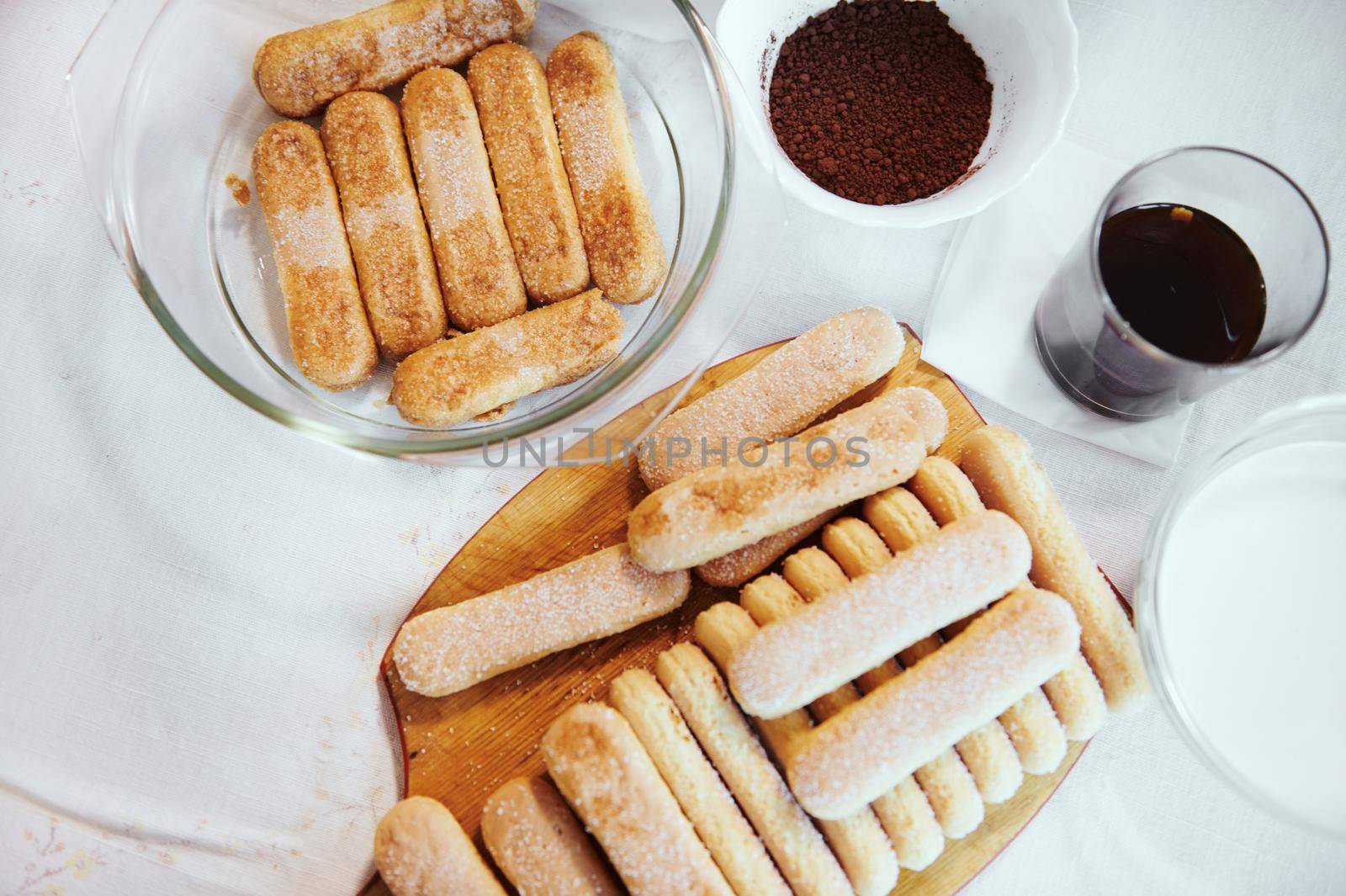 Flat lay. Ingredients for making traditional Italian tiramisu on kitchen table - savoiardi biscuit cookies on wooden board, sweet espresso coffee in glass, mascarpone cheese and bowl with fresh cream