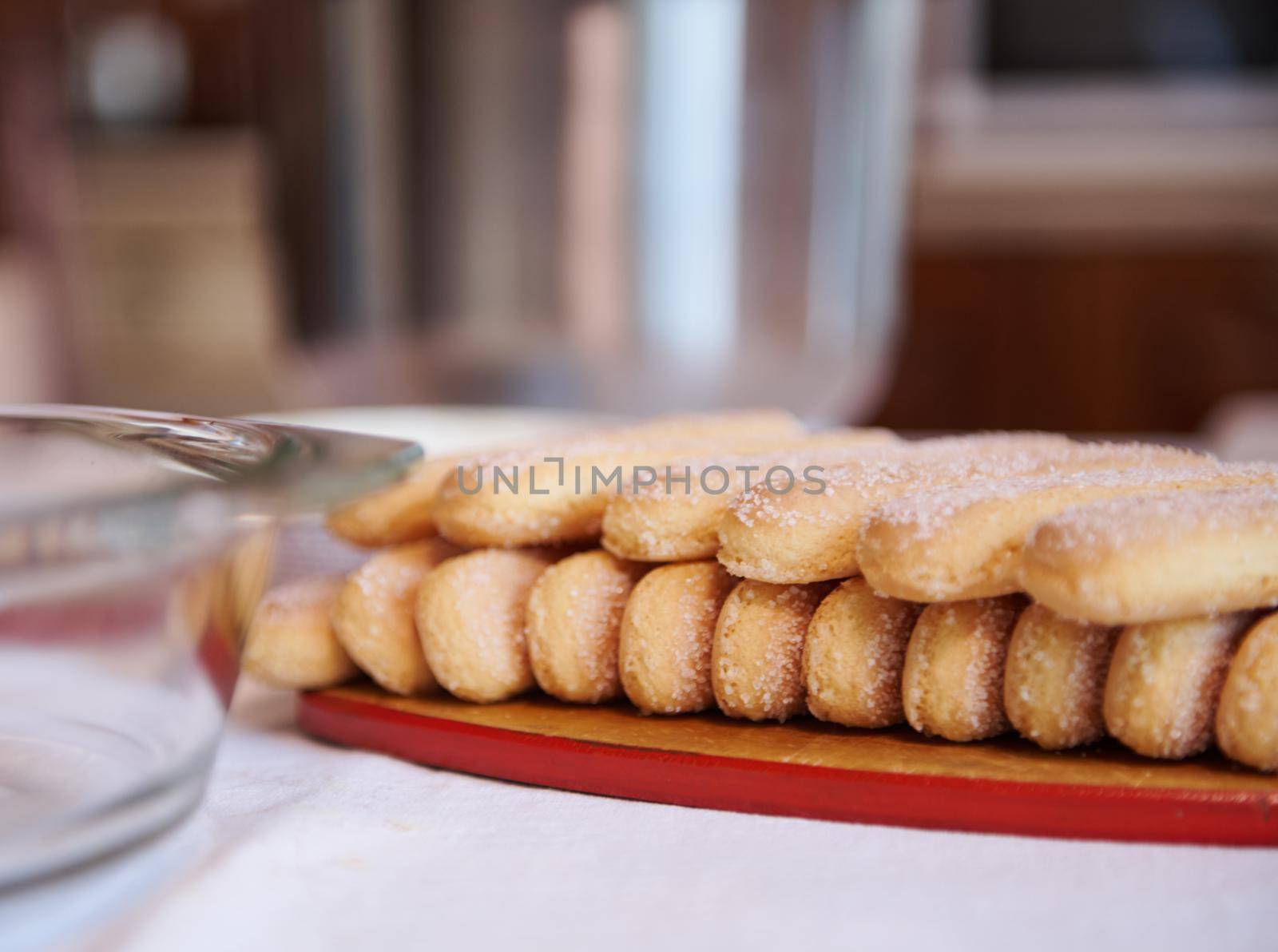 Close-up of ladyfinger savoiardi biscuit cookies on a wooden cutting board on white tablecloth background with blurred steel bowl and glass plate. Close up