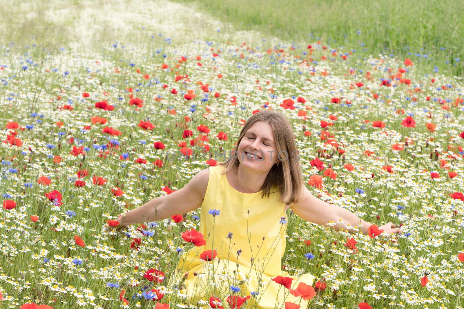 a beautiful young blonde woman in a yellow dress stands among a flowering field of poppies, daisies, cornflowers and laughs. High quality photo