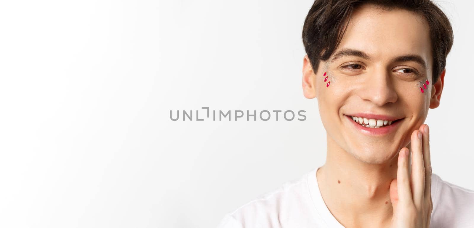 People, lgbtq and beauty concept. Headshot of beautiful gay man with glitter on face, smiling and looking happy, touching cheek after kiss, white background by Benzoix