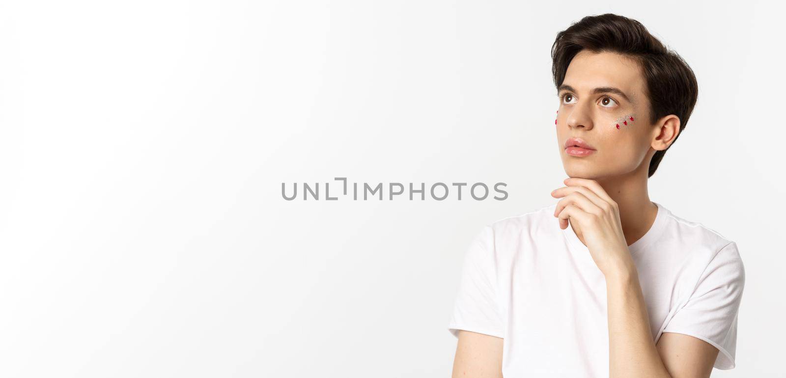People, lgbtq community and lifestyle concept. Beautiful androgynous man with glitter on face, looking at upper left corner sensual, standing over white background.