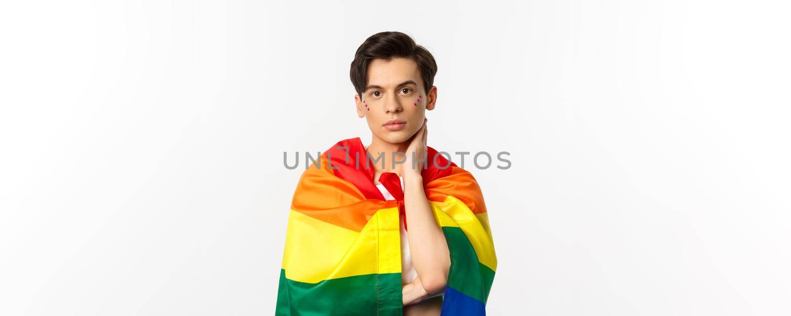 Waist-up shot of beautiful gay man with glitter on face, wrap himself with rainbow lgbt flag and touching face gently, looking at camera, white background.