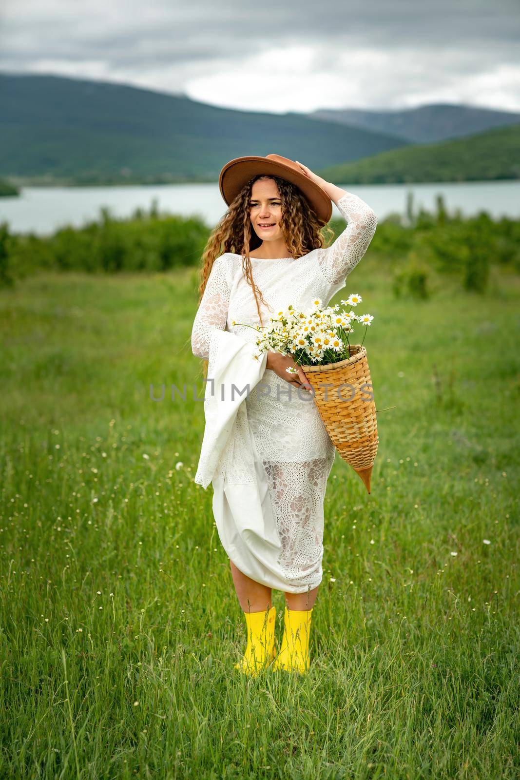 A middle-aged woman in a white dress and brown hat holds a lA middle-aged woman in a white dress and brown hat holds a basket in her hands with a large bouquet of daisies.arge bouquet of daisies in her hands. Wildflowers for congratulations by Matiunina