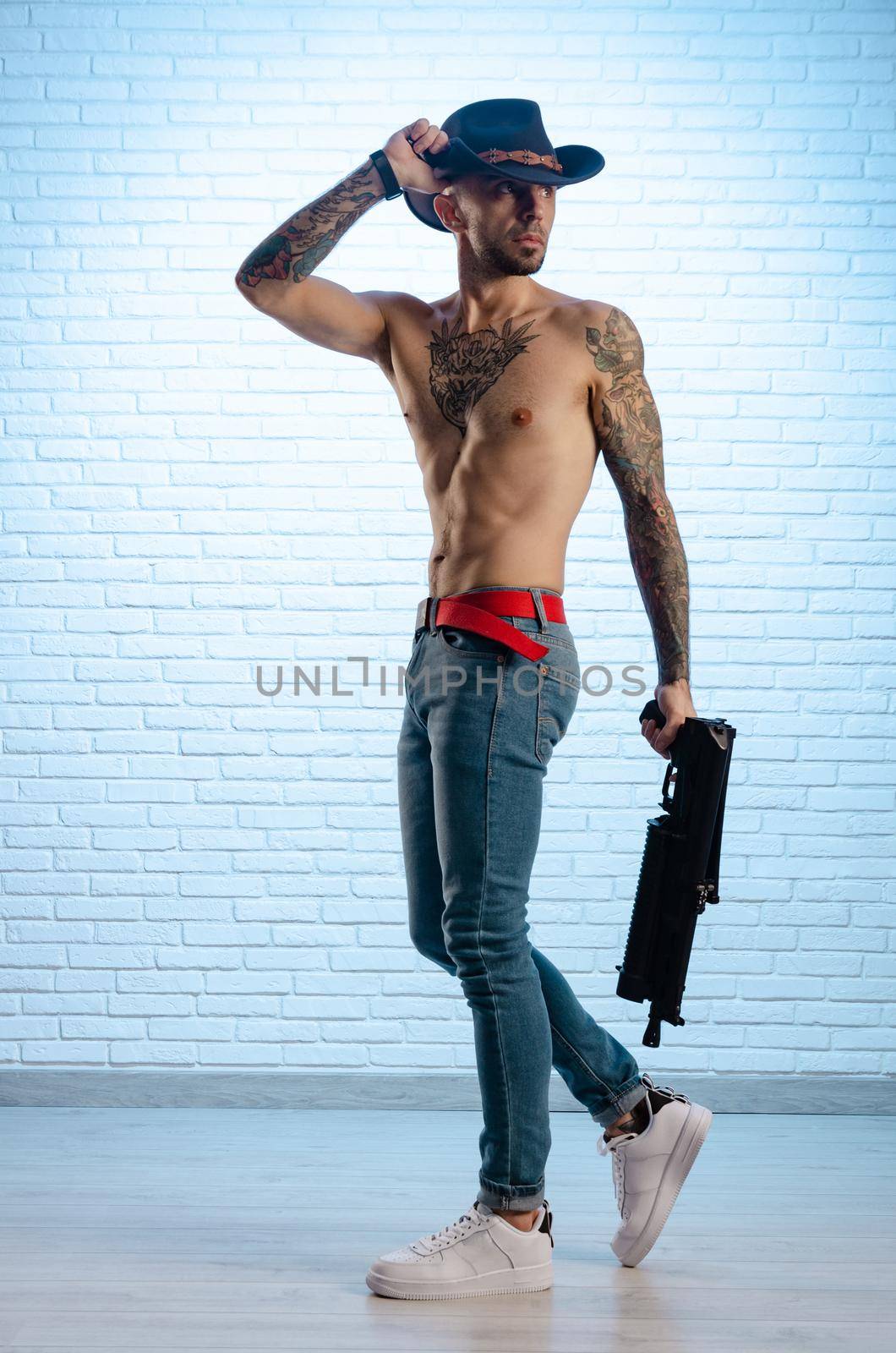 the fashionable slim guy with a bare torso in tattoos, jeans and a cowboy hat with a gun