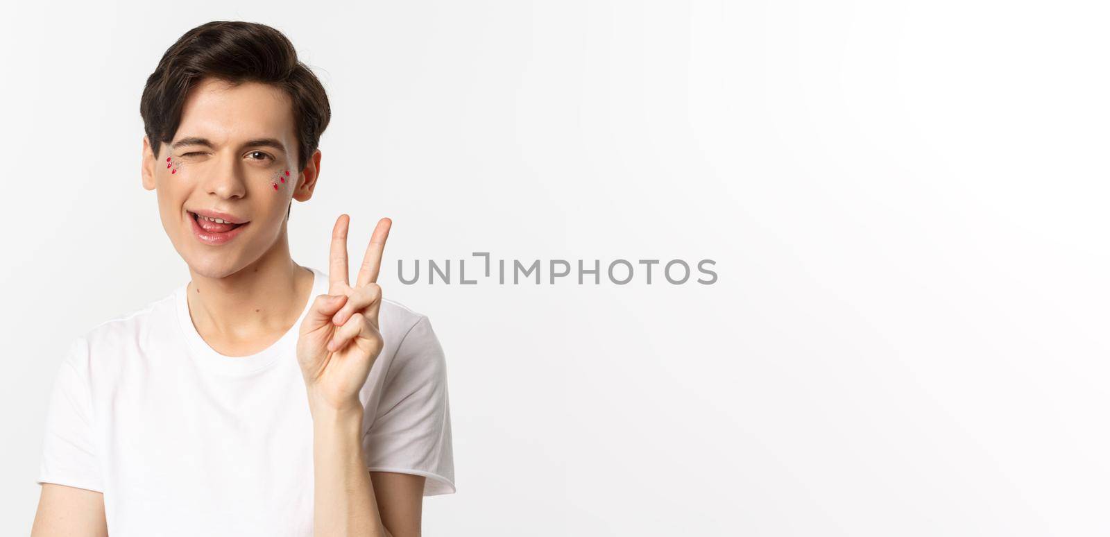 People, lgbtq community and lifestyle concept. Happy and cute gay man with glitter on face, showing peace sign and smiling, celebrating pride holiday, white background by Benzoix