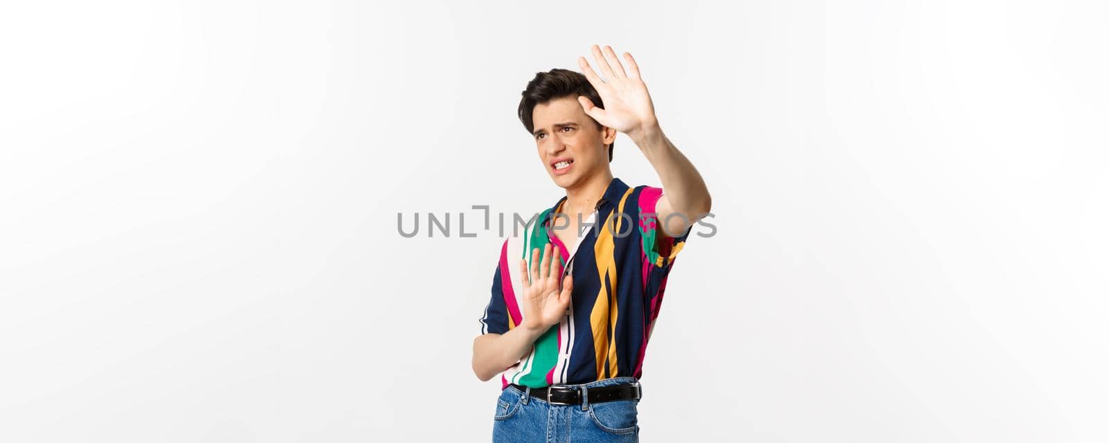Timid and displeased androgynous man asking to stop, raising hands defensive and grimacing, standing over white background.