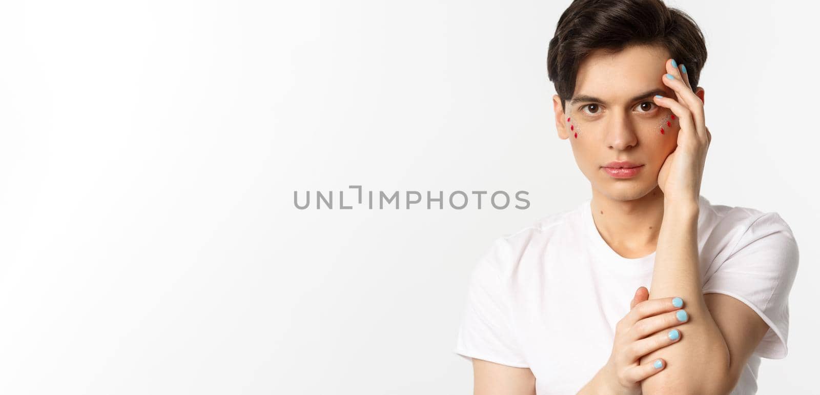 People, lgbtq and beauty concept. Close-up of beautiful queer man touching face with fingers with blue nail polish, standing over white background by Benzoix