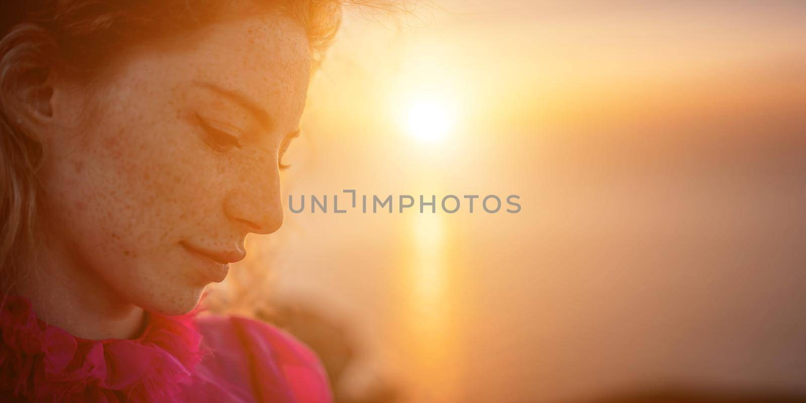 Side view a Young beautiful sensual woman in a red long dress posing on a volcanic rock high above the sea during sunset. Girl on the nature on overcast sky background. Fashion photo