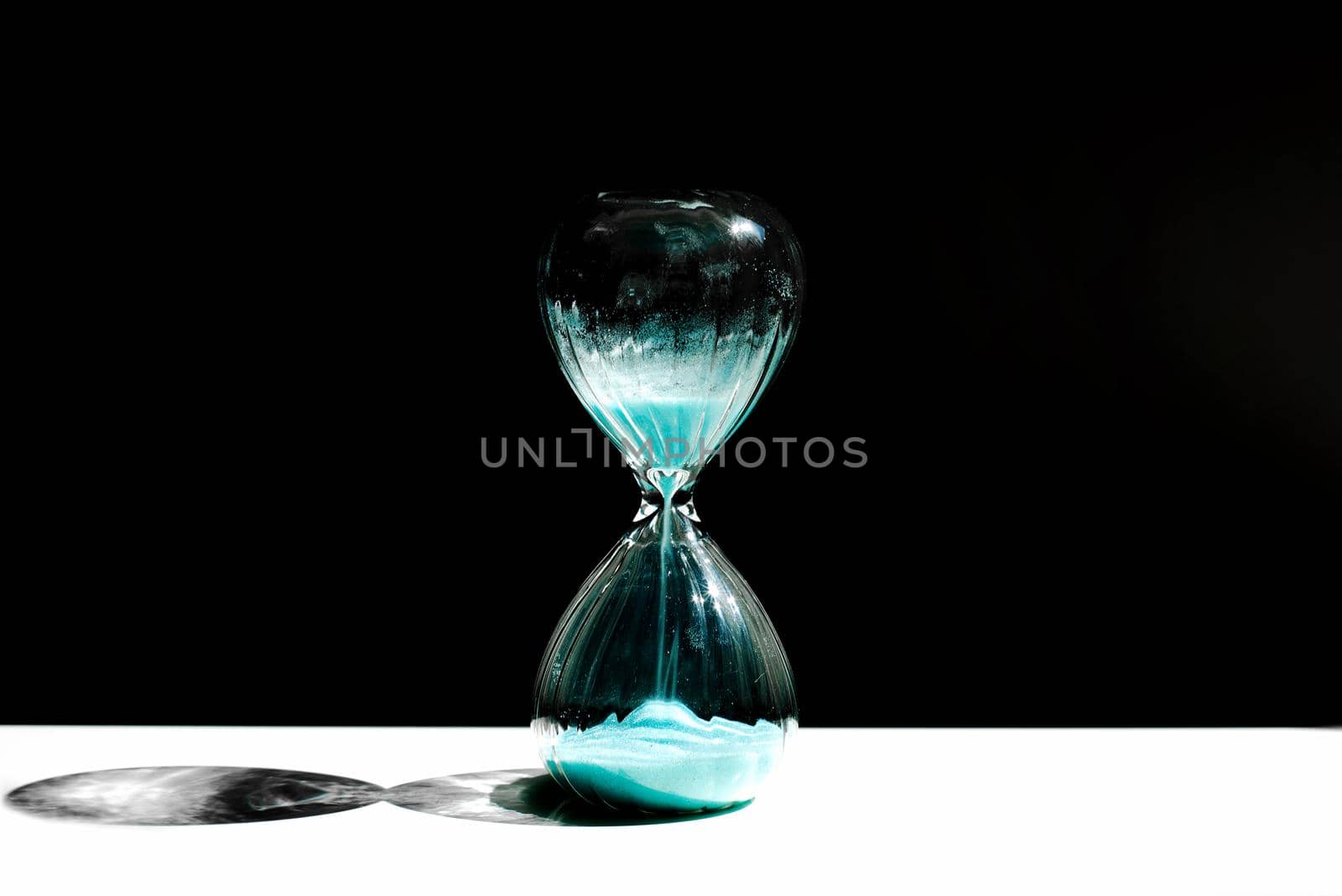 Hourglass, hourglass with blue sand on a black background with a streak of sunlight and a shadow