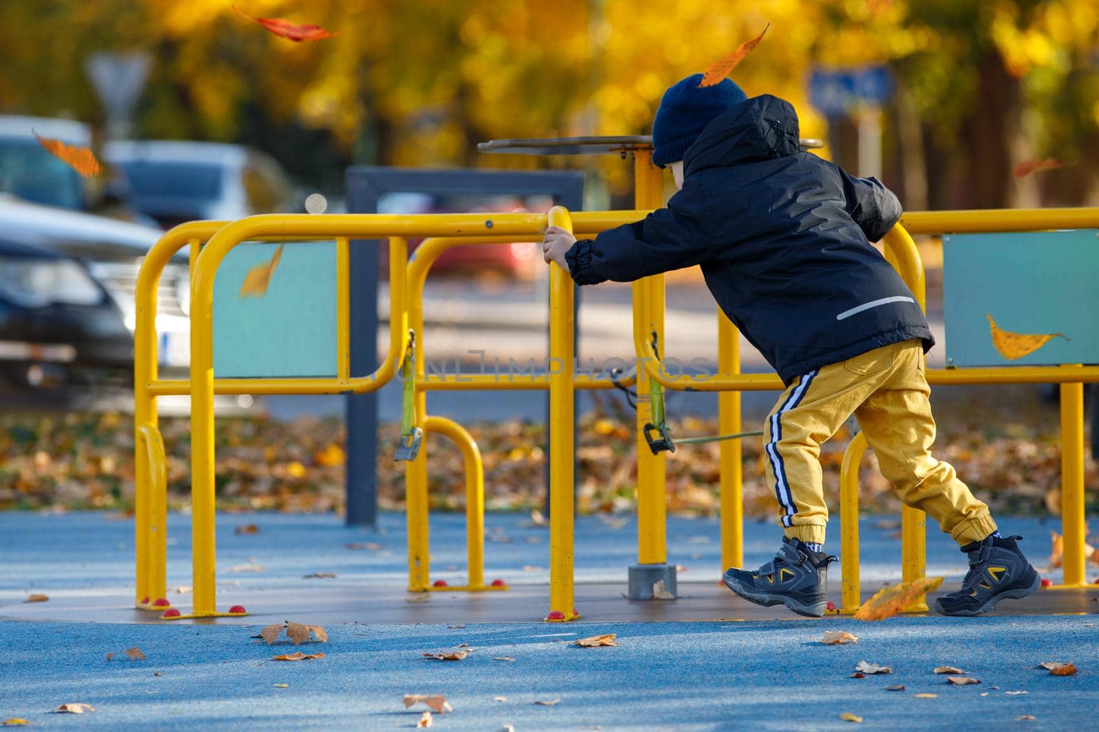 Little boy play on the playground in the park. Child spinning a yellow carousel. Autumn season with bright yellow trees in the background. Vertical photo.