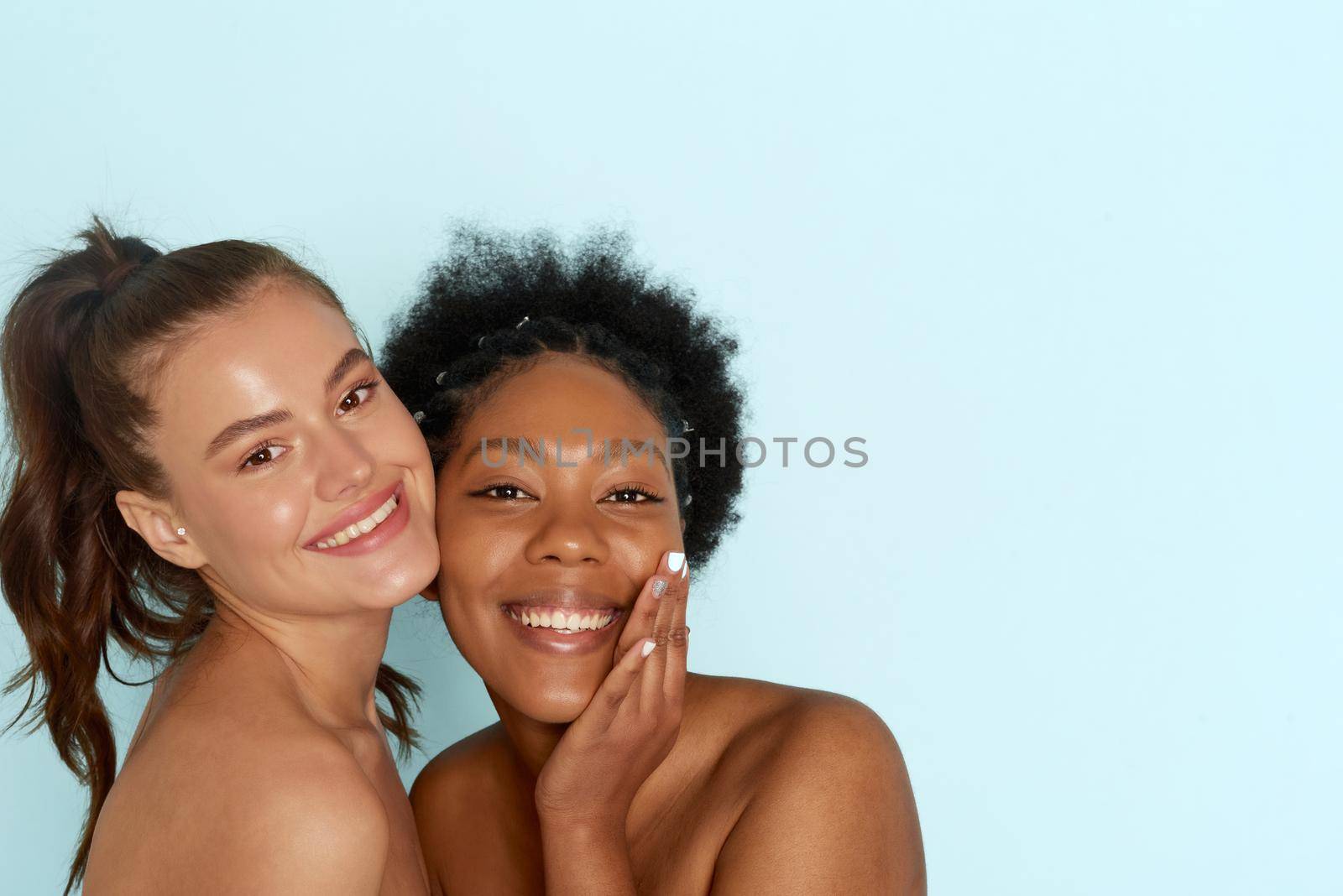 Portrait of two young multiracial women with perfect moisturizing skin standing together and smiling at the camera against a blue background. The concept of different cultures, care for the skin, hair