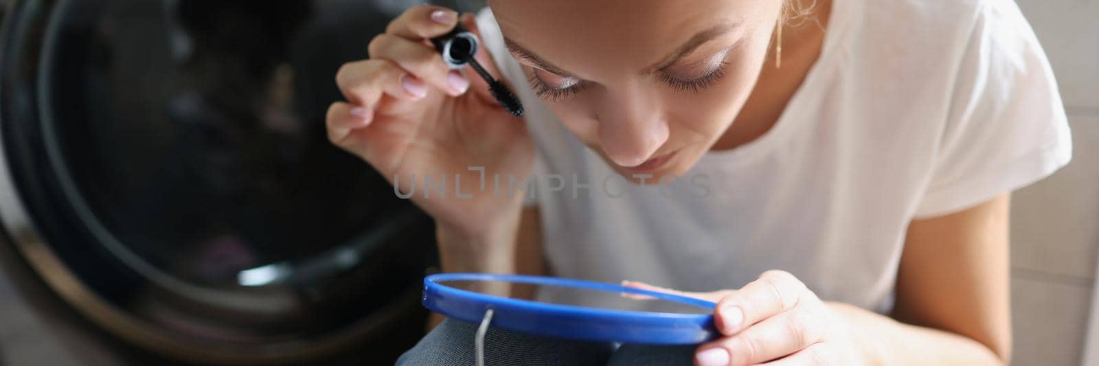 Close-up of woman getting ready for work, applying mascara using small mirror. Female in rush, doing makeup on floor in bathroom. Get ready, hurry, morning routine concept