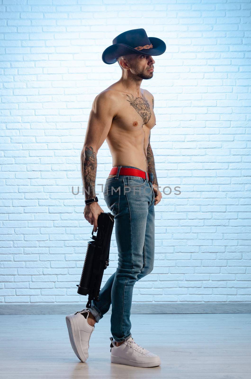 the fashionable slim guy with a bare torso in tattoos, jeans and a cowboy hat with a gun