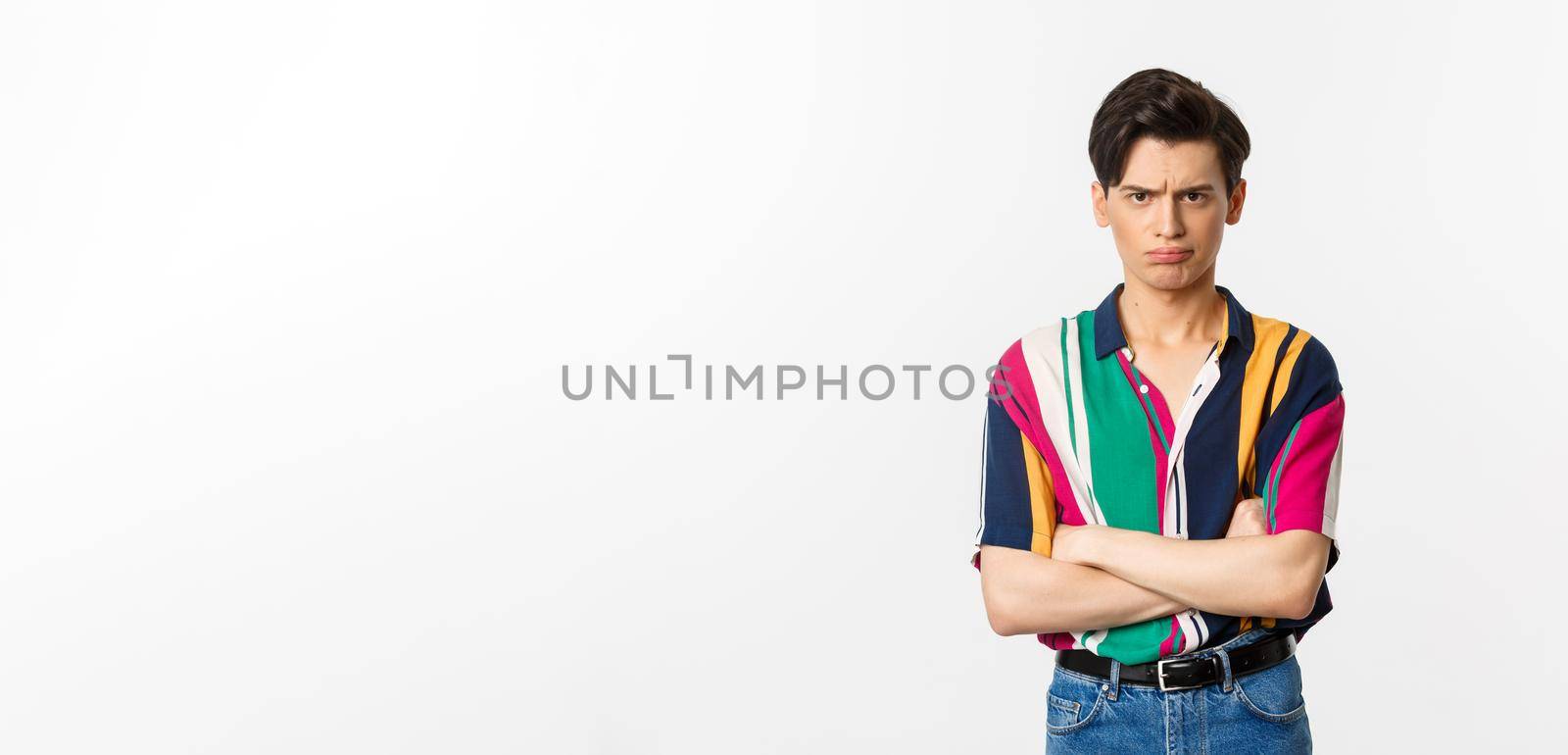 Offended and angry young gay man sulking, cross arms chest and looking judgemental at camera, standing over white background by Benzoix