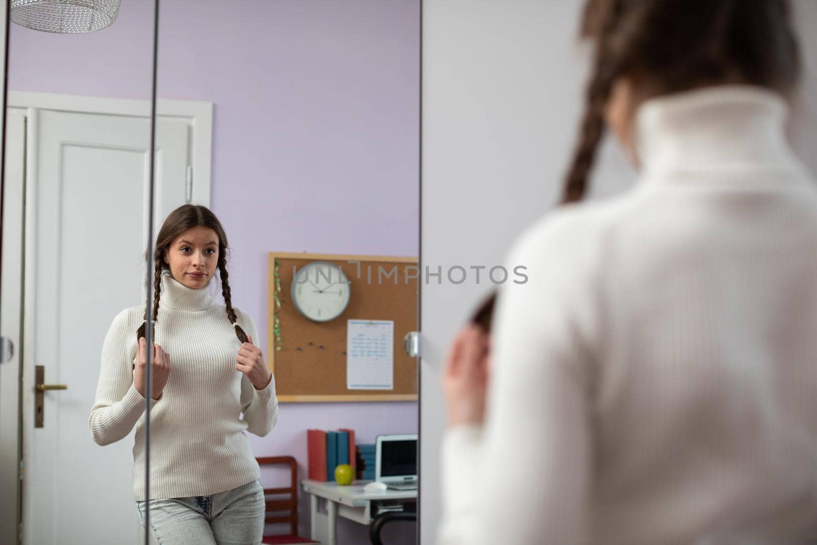 Young brunette girl poses in front of large mirror in room and changes facial expressions. Attractive young girl with two braids.