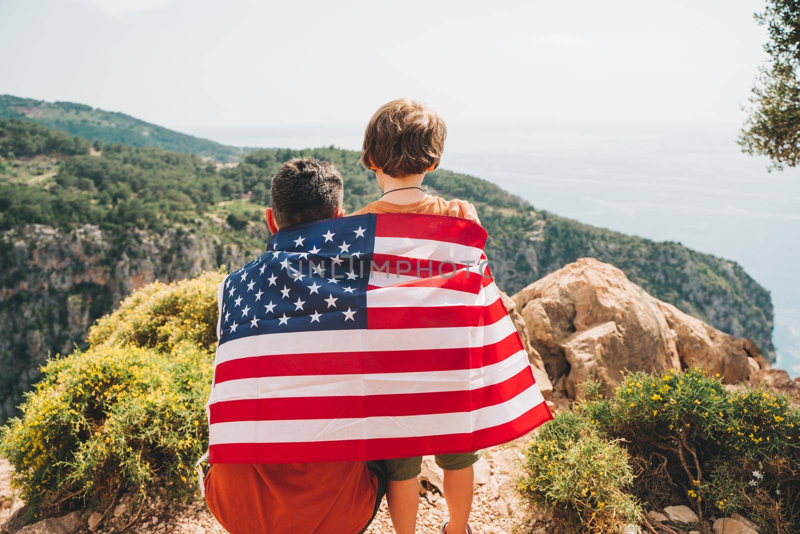 Young man dad father and his son school boy kid standing on a rock cliff with US flag on shoulders and looking at sea. Travellers child wearing American flag on back. 4 July Independence Day