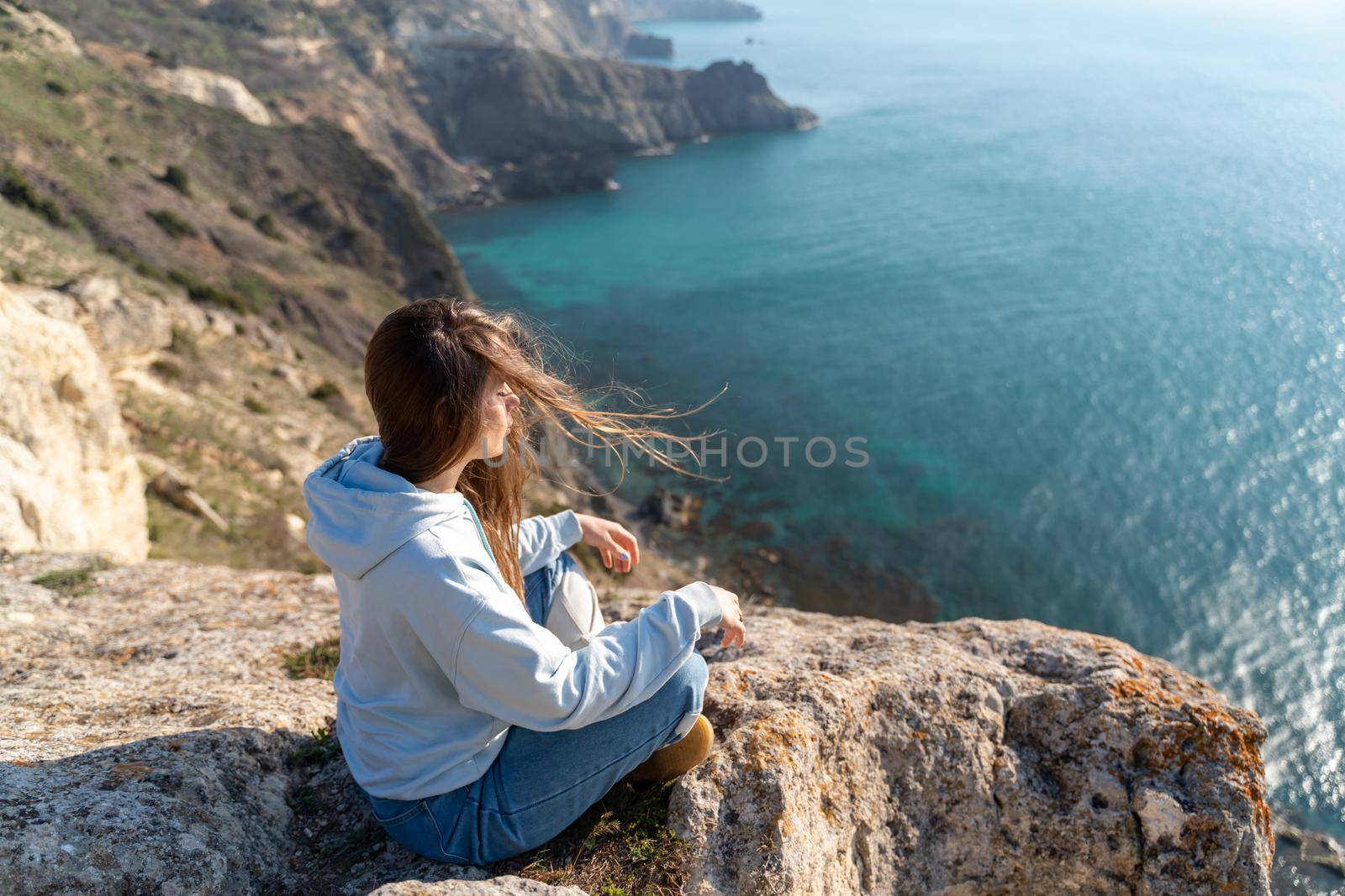 Woman tourist enjoying the sunset over the sea mountain landscape. Sits outdoors on a rock above the sea. She is wearing jeans and a blue hoodie. Healthy lifestyle, harmony and meditation.